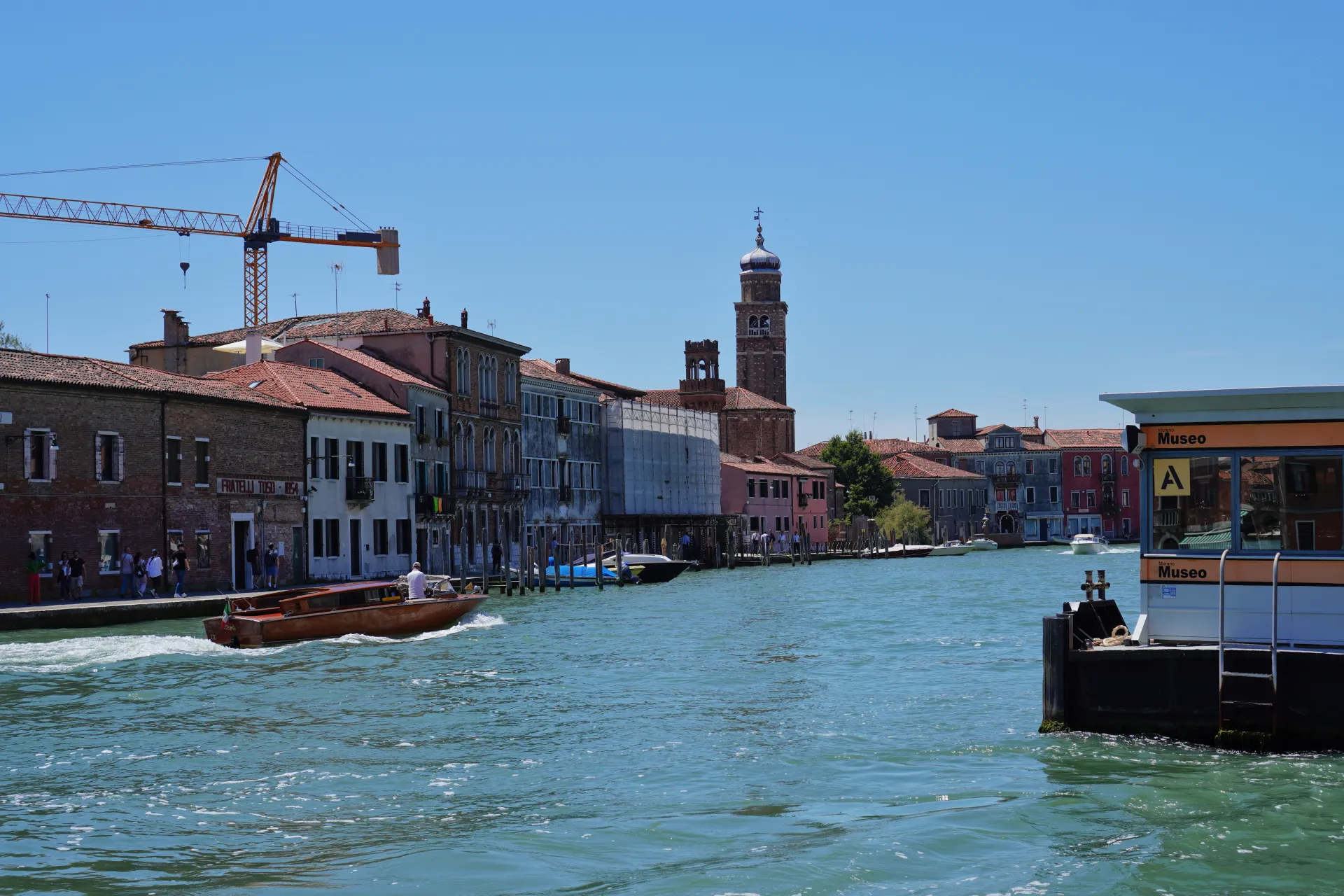 A scenic canal in Venice, Italy, with a water taxi cruising along. The buildings lining the canal showcase traditional Venetian style. In the distance, the prominent bell tower of Murano Island stands out against the clear blue sky.