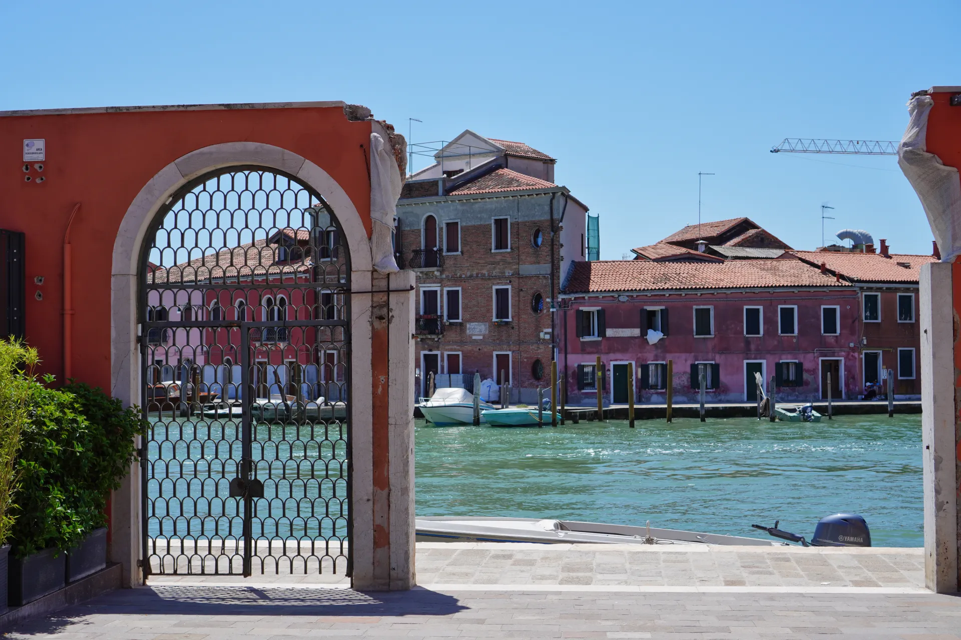 View through an ornate metal gate of a canal on Murano island, Venice, with colorful Venetian buildings and boats.