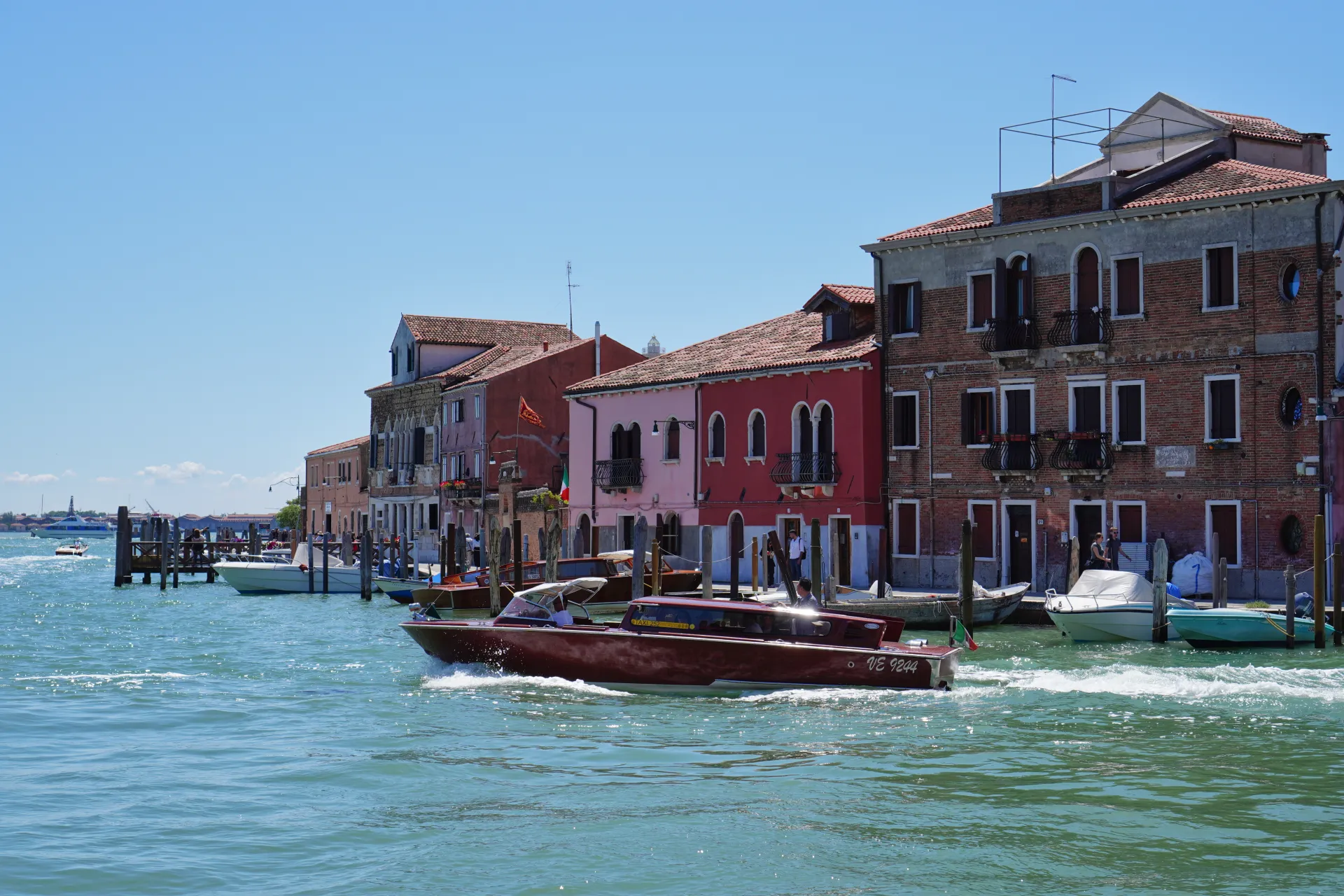 Čamac plovi kanalom duž koga se nižu šarene zgrade na ostrvu Murano, Venecija, Italija, pod vedrim plavim nebom.