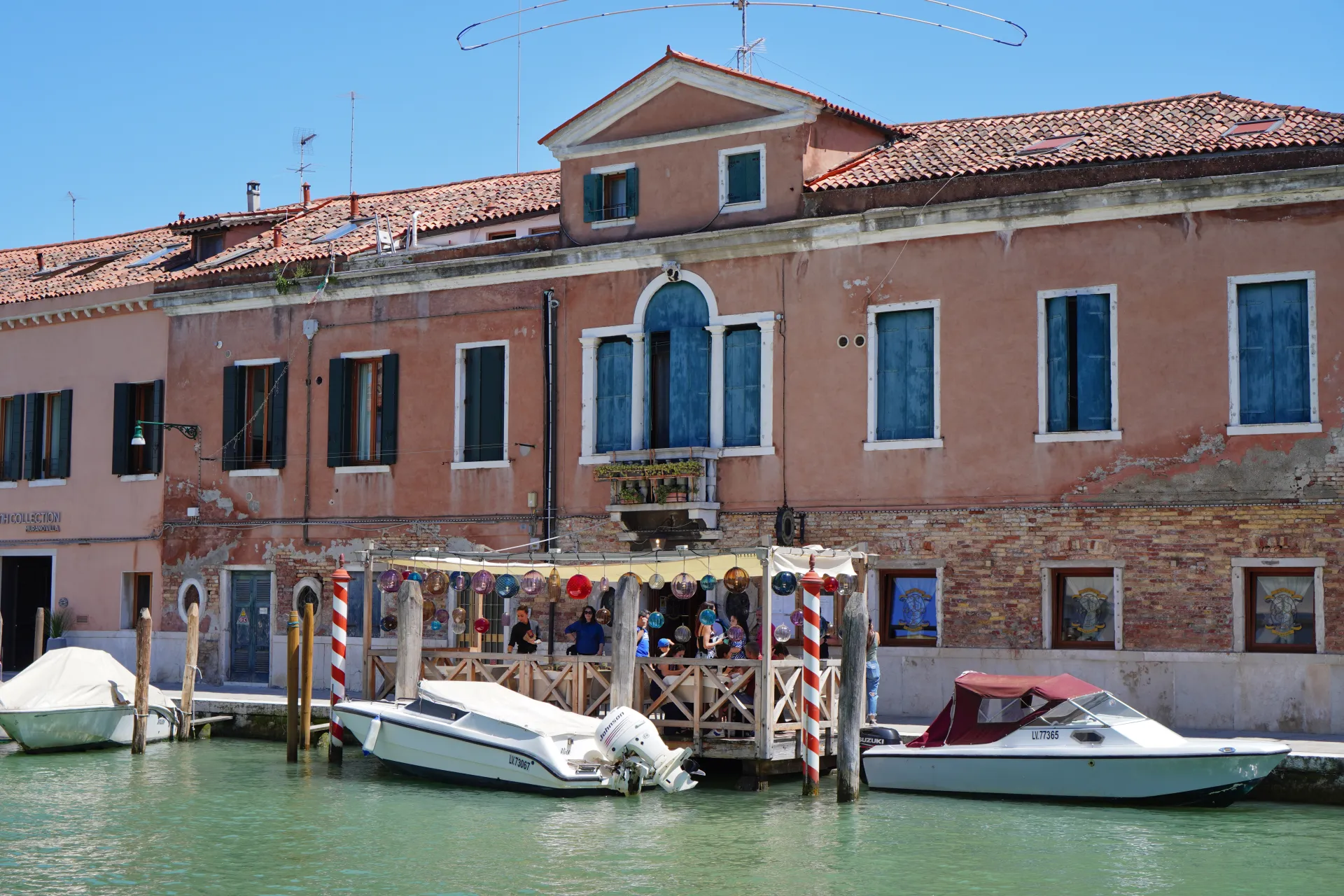 A picturesque canal lined with buildings on Murano Island, Venice, Italy. Boats are docked, and colorful glass orbs hang as decorations above a waterfront area.