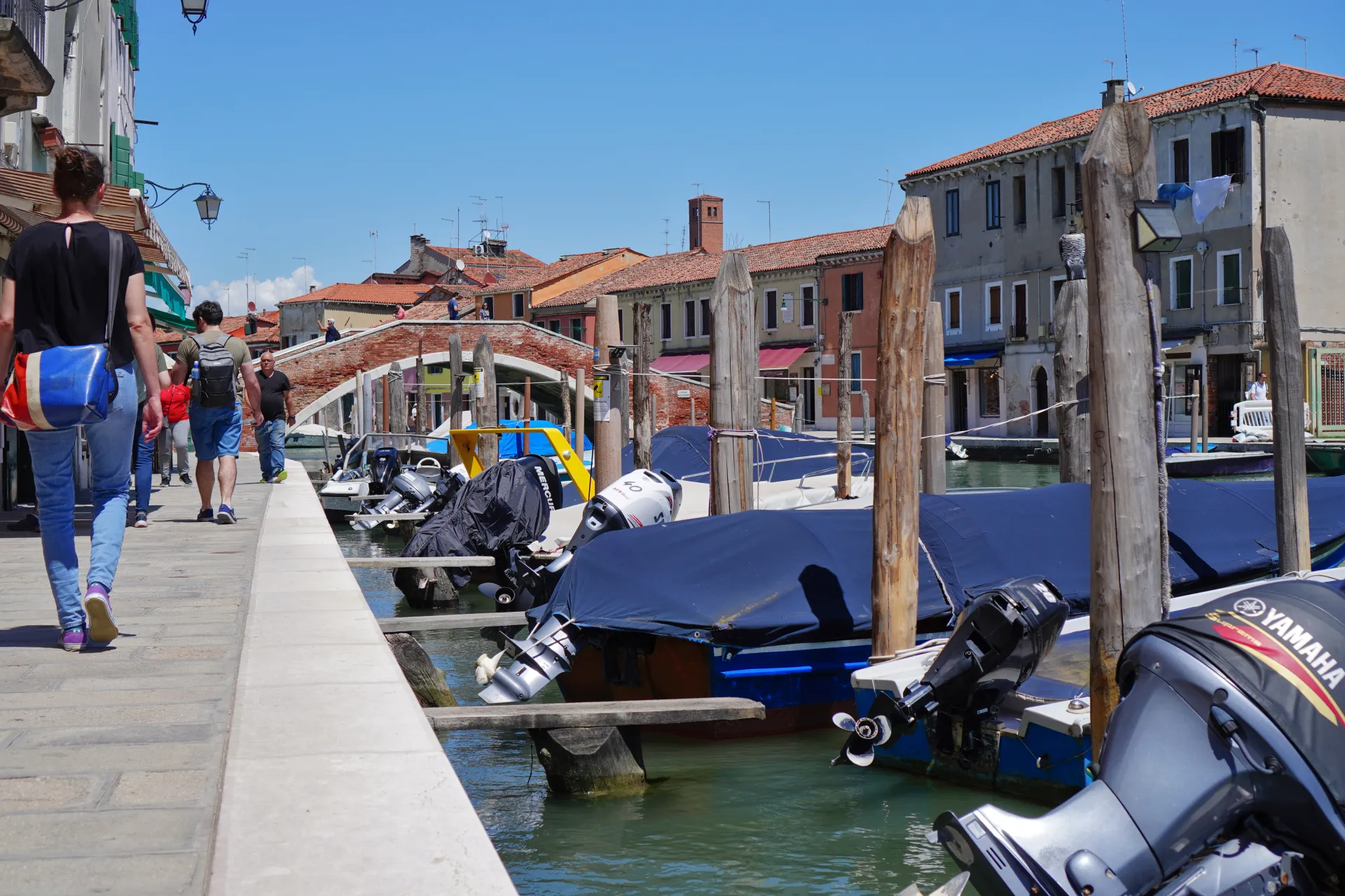 People walking along a canal on Murano island in Venice, with boats docked and buildings in the background.