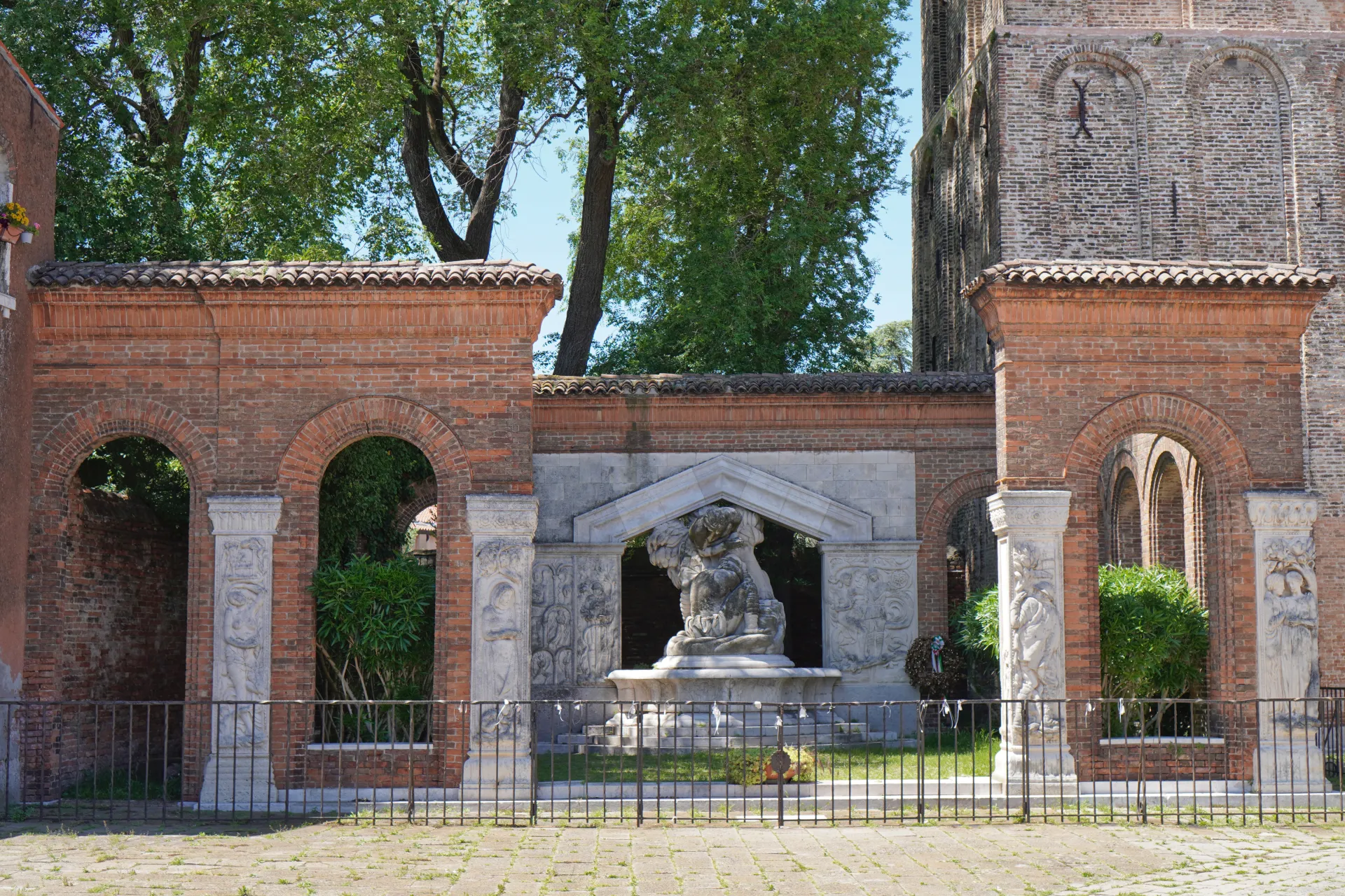 A weathered stone monumental fountain with sculpted figures and reliefs, set within arched brick structures on Murano island in Venice, Italy. Green trees and a tall church tower are visible behind the fountain.