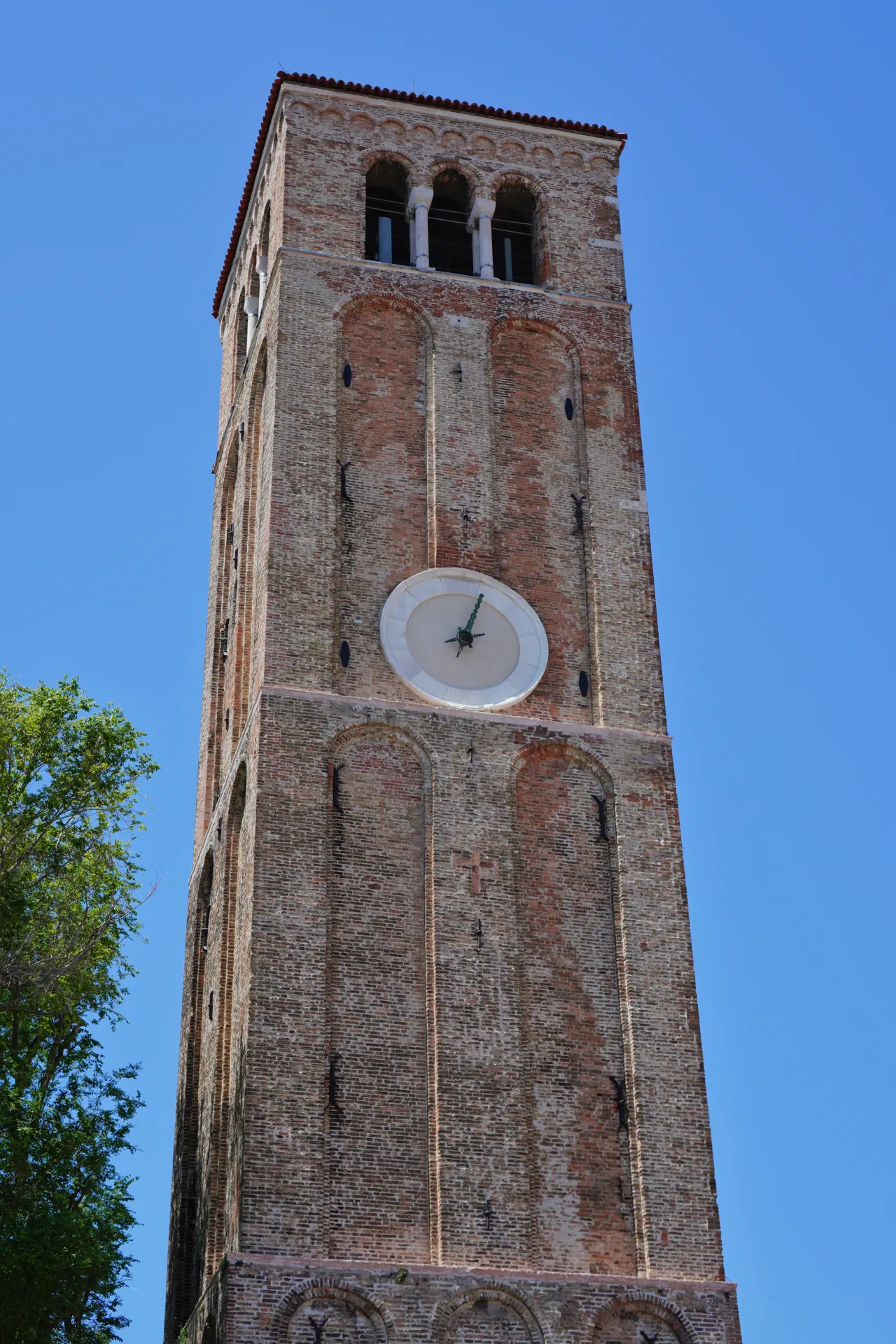 The brick clock tower on Murano Island in Venice, Italy, stands tall against a bright blue sky, with a clock face and arched openings.