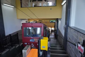 A red funicular train is parked at a station with a set of stairs going up to the exit and ticket barriers in the foreground.