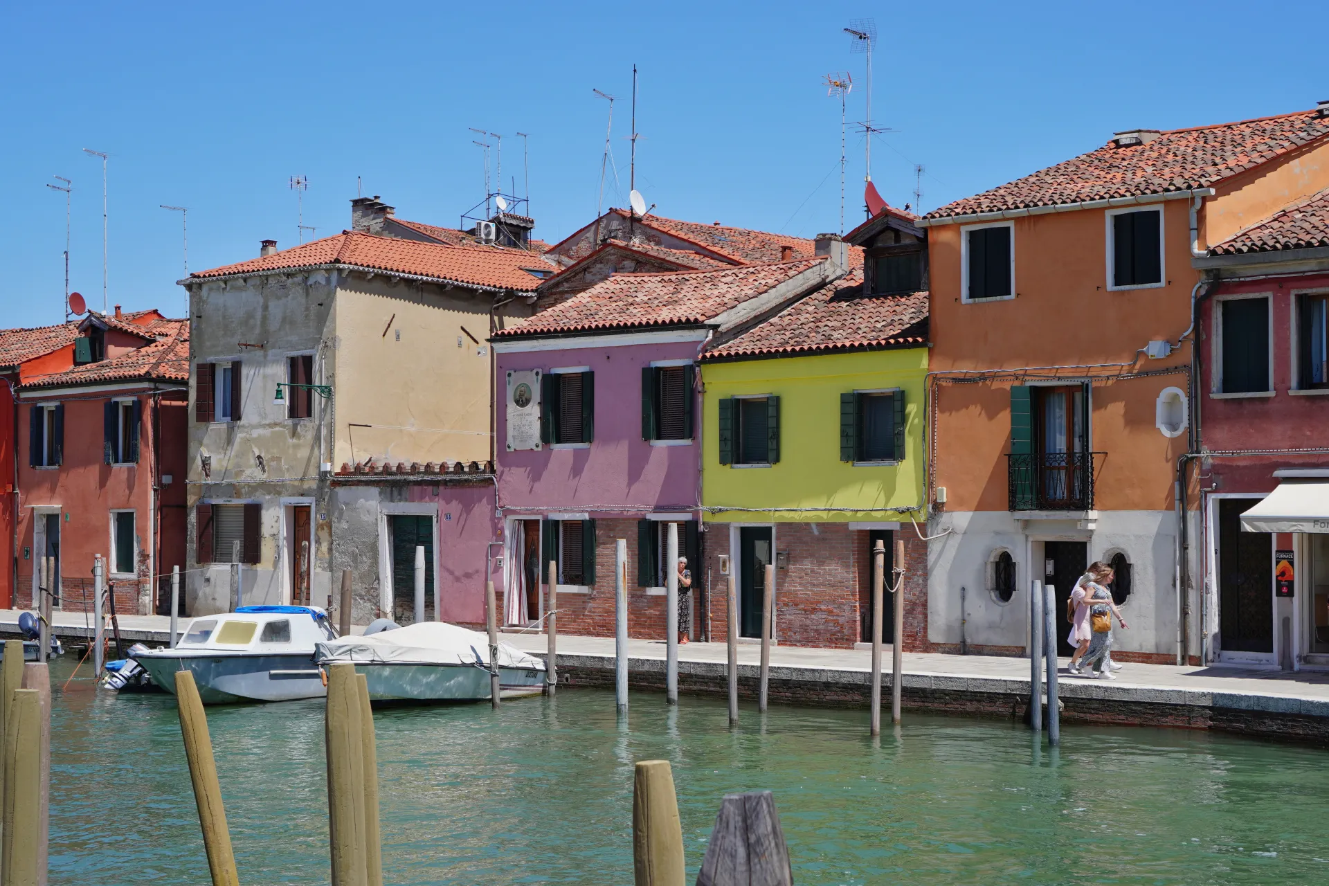 Colorful houses in shades of pink, yellow, and orange stand along a canal on Murano island, Venice, with boats moored to wooden posts and people walking on the waterfront promenade.
