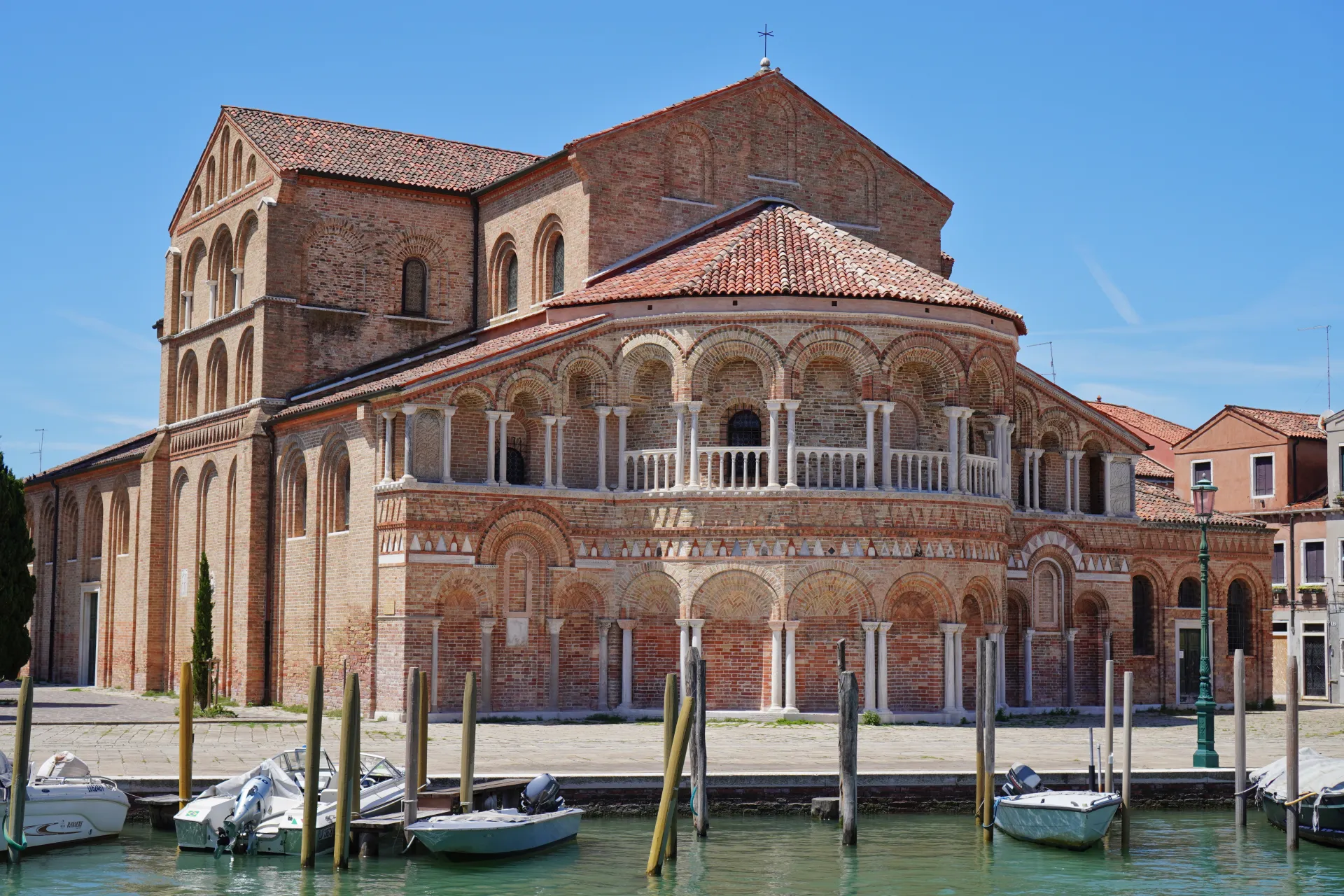 Exterior view of the Basilica di San Donato, a large brick church with arched windows and a tiled roof, situated on the canal on the island of Murano in Venice. Several boats are docked in the turquoise water in front of the church.