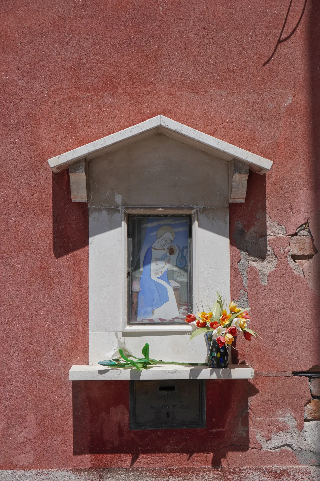 Svetište sa slikom Madone sa Detetom, ukrašeno cvećem, na teksturiranom crvenom zidu na ostrvu Murano, Venecija, Italija.