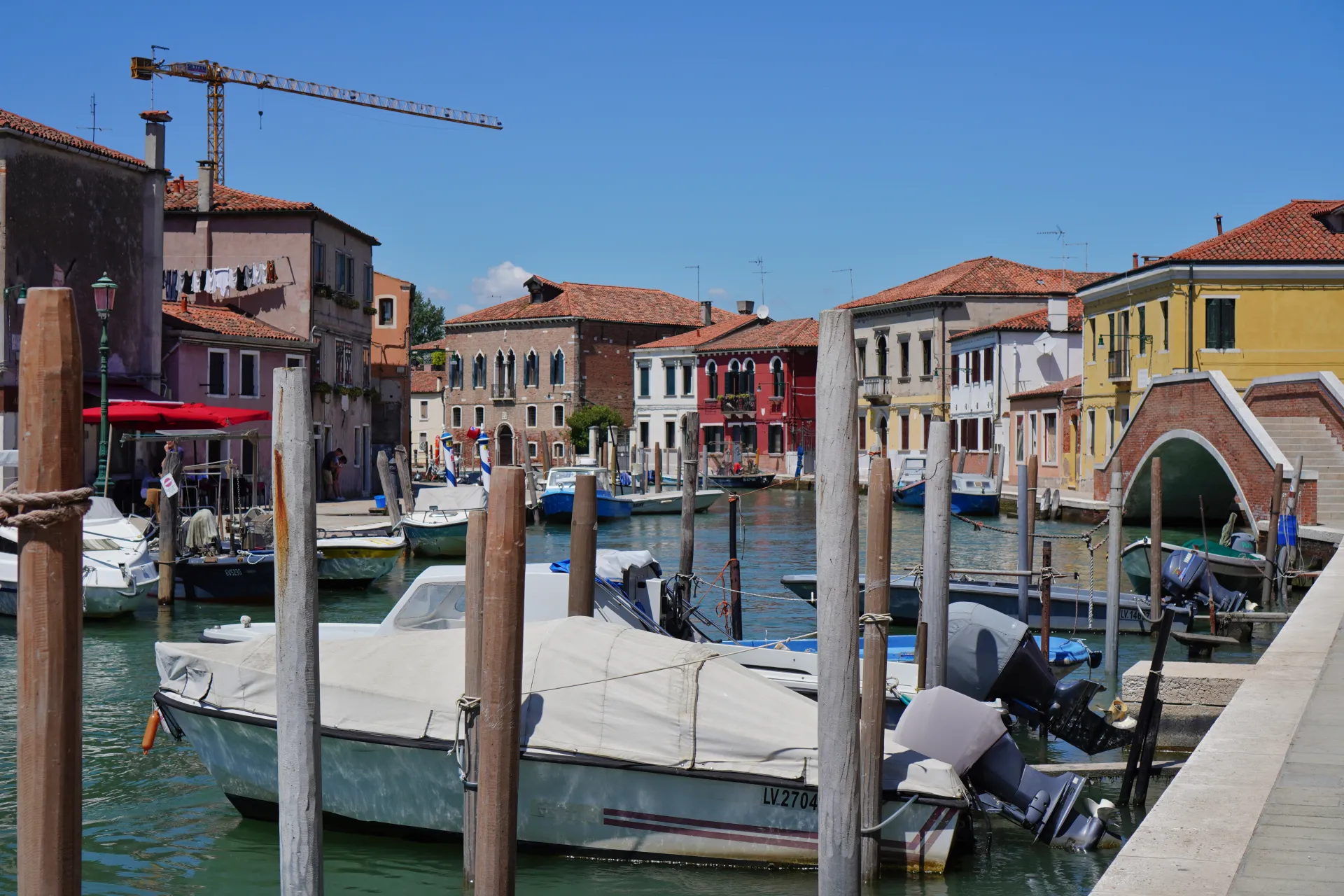 A canal on Murano island in Venice, Italy, with several boats tied to wooden posts. Buildings with terracotta roofs and colorful facades line the canal. Laundry is hanging from a clothesline on a building, and a construction crane is visible in the background under a clear blue sky.