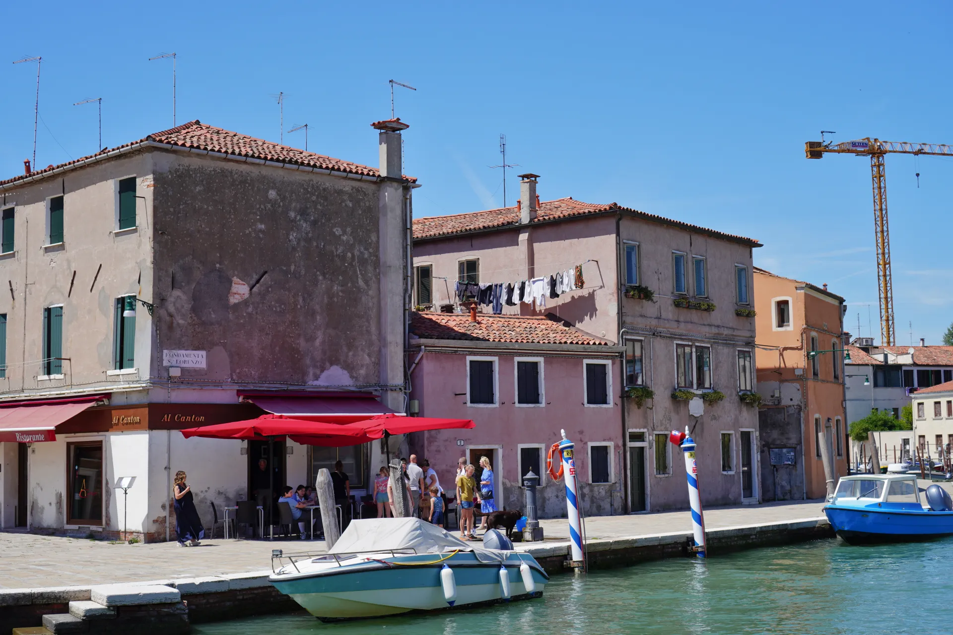 A sunny day scene on Murano Island, Venice, Italy, showing buildings along a canal, a cafe with red umbrellas, boats docked, and laundry hanging out to dry. A construction crane is visible in the background.