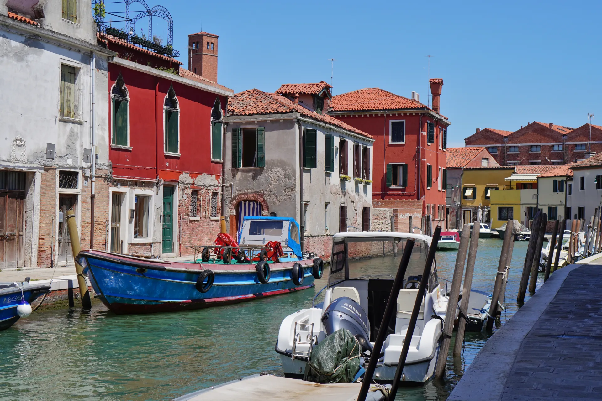 A bright blue boat and modern motorboat are moored in a canal on Murano island, Venice. Colorful buildings line the waterway, under a clear blue sky.