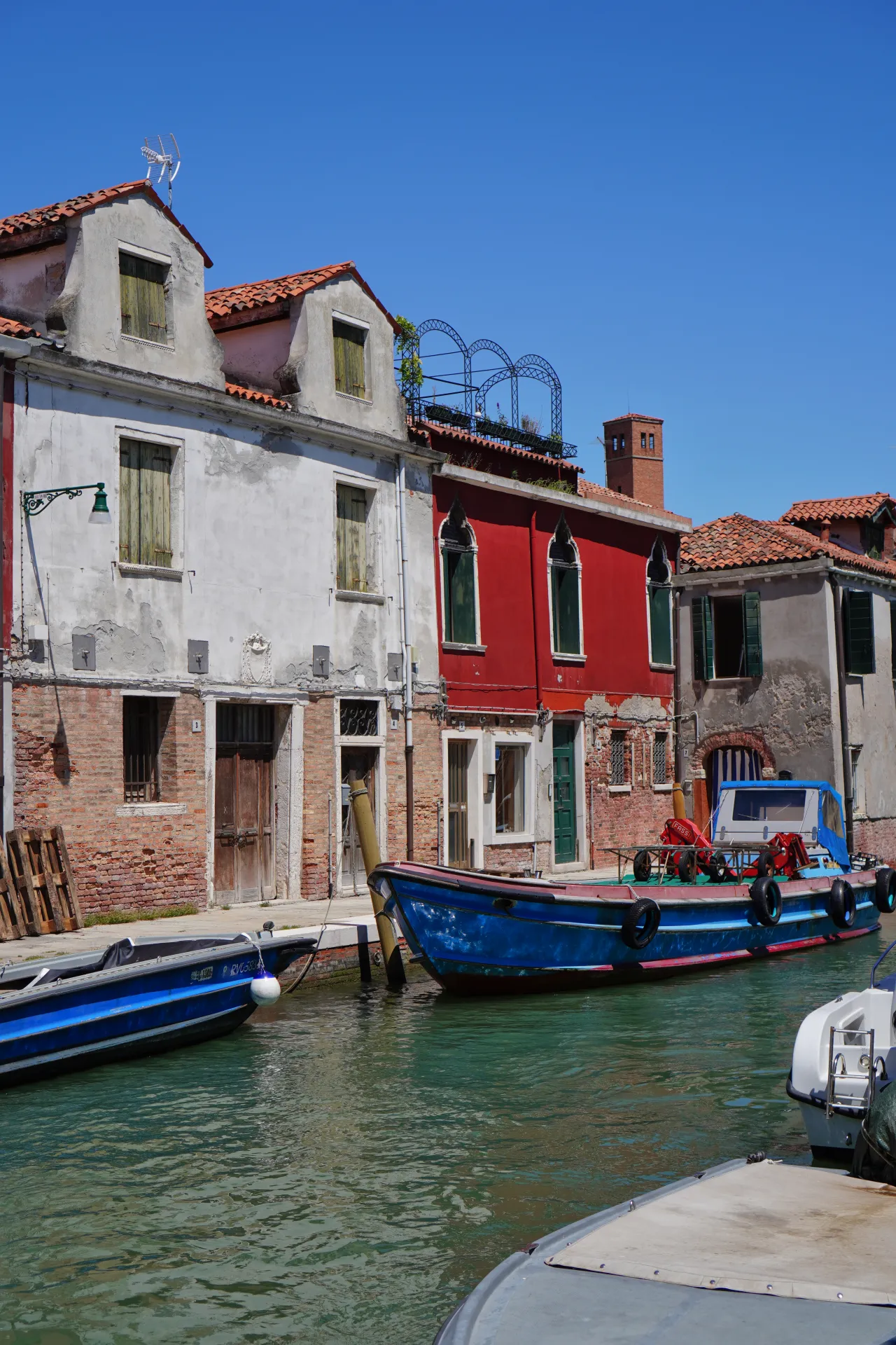 A canal lined with traditional Venetian houses, some with weathered facades and green shutters, and a bright red building, on Murano island in Venice, Italy. Several boats are docked in the turquoise canal, reflecting the clear blue sky.