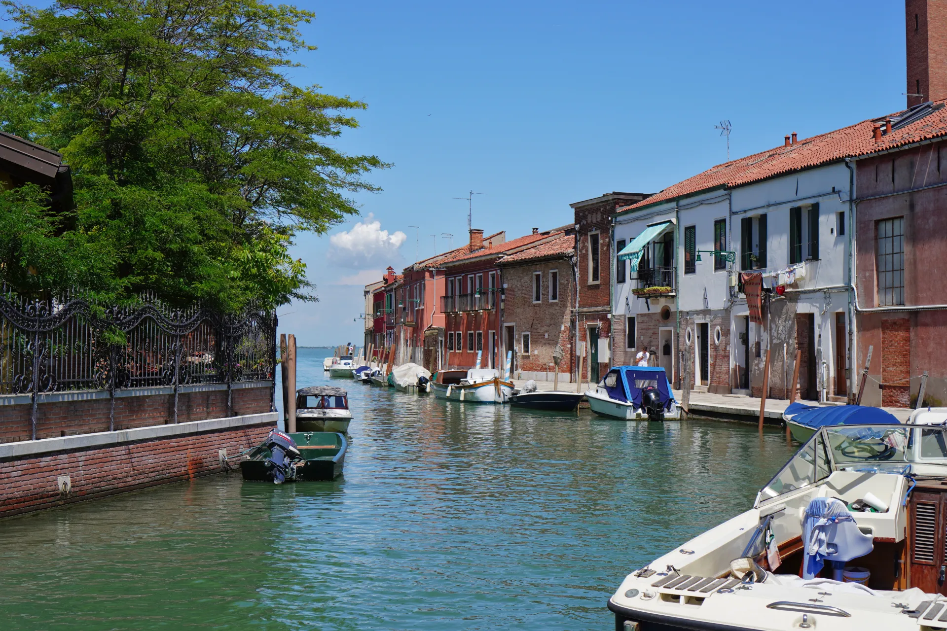 A tranquil canal on the island of Murano in Venice, Italy, with colorful houses lining the banks and several boats moored in the water. A large green tree provides shade on the left side of the image.