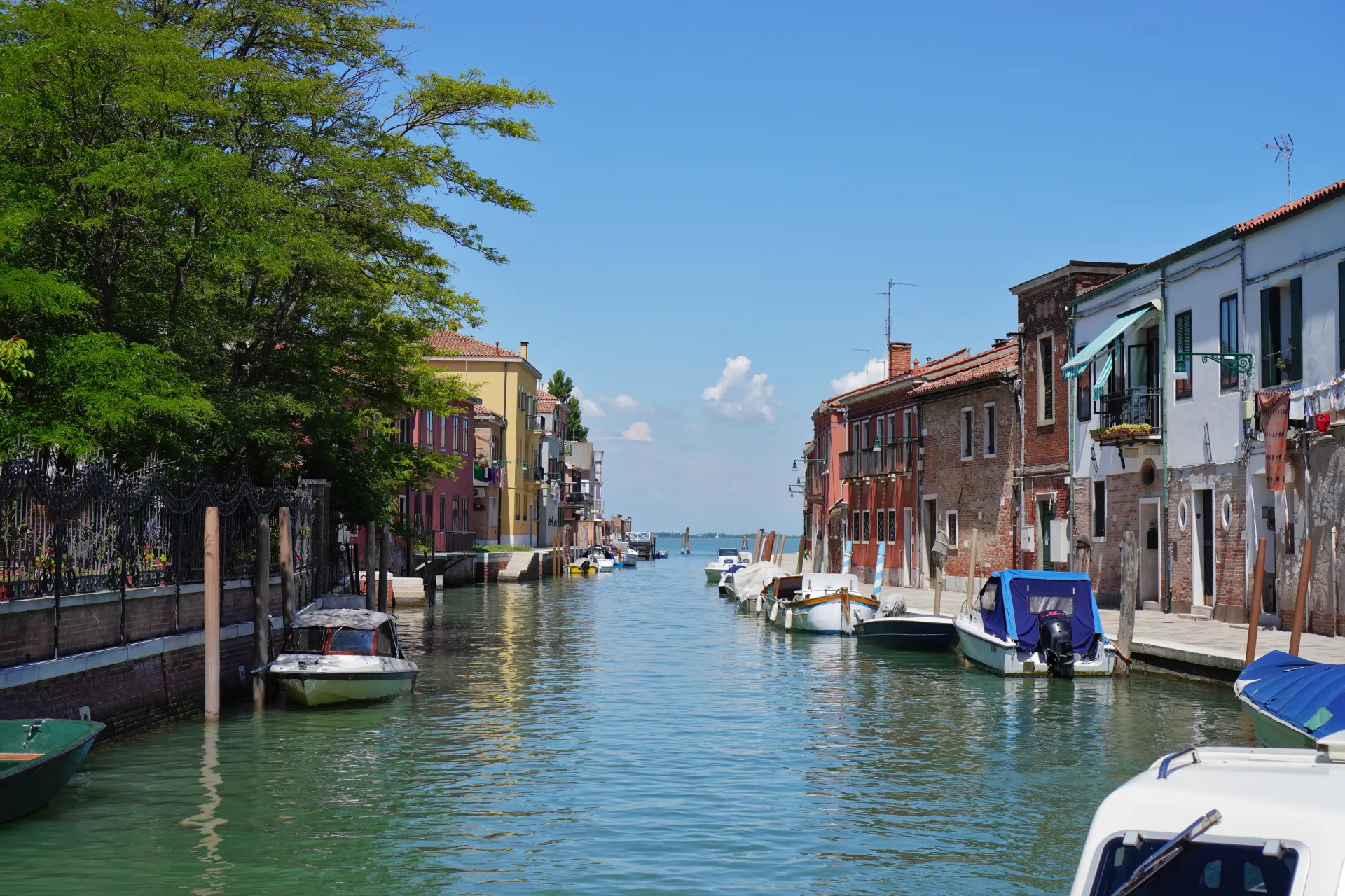 A peaceful canal in Murano, Venice, Italy, with colorful houses, moored boats, and trees reflecting in the water.