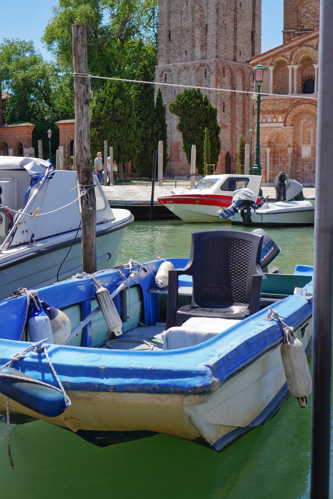 Several boats are docked in a canal on Murano Island, Venice, with traditional brick buildings and trees in the background.