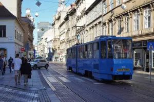 A bright blue tram travels down the tram tracks on Ilica Street in Zagreb, Croatia. Pedestrians walk on the sidewalk beside ornate buildings, and a white car is stopped on the tracks.
