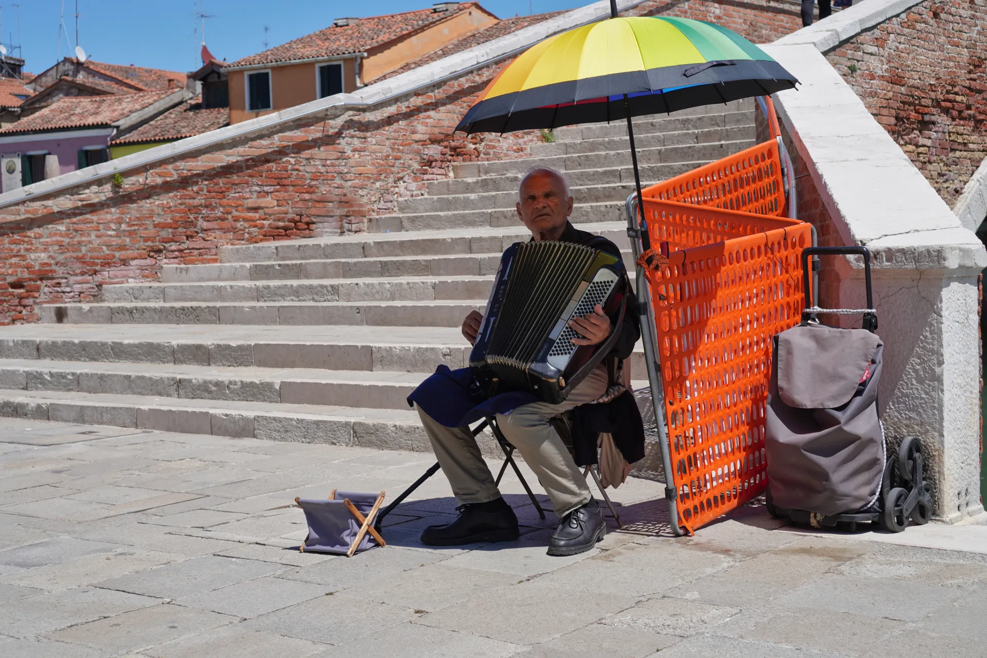 Stari čovek svira harmoniku na mostu na ostrvu Murano, Venecija, sa kišobranom duginih boja iznad njega i stepenicama u pozadini.