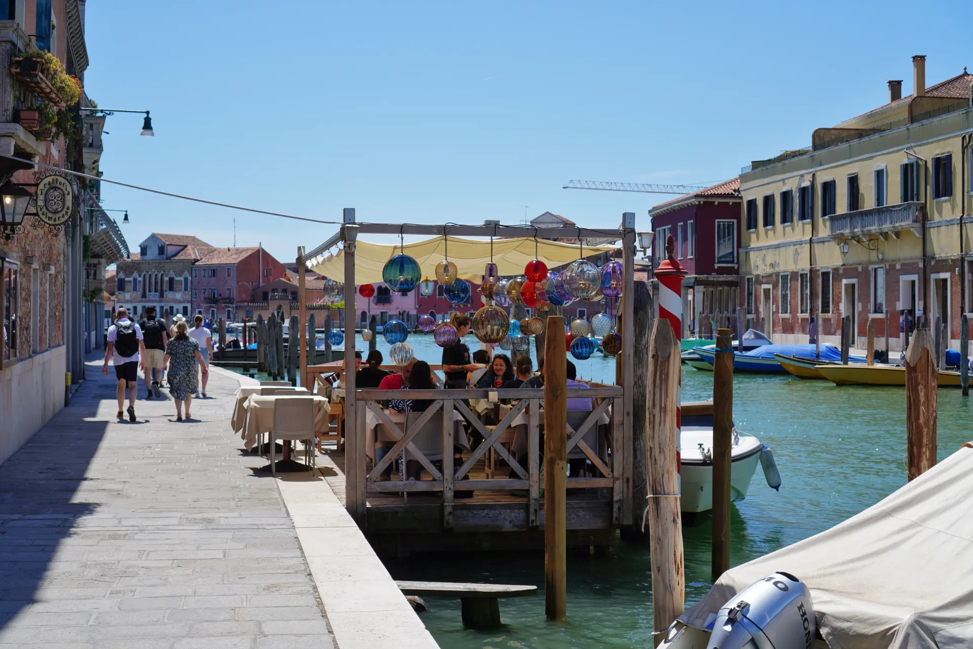 People dining at a waterfront restaurant in Venice, with colorful Murano glass spheres hanging from the canopy, overlooking a canal with boats. The island of Murano is famous for its glassmaking tradition.