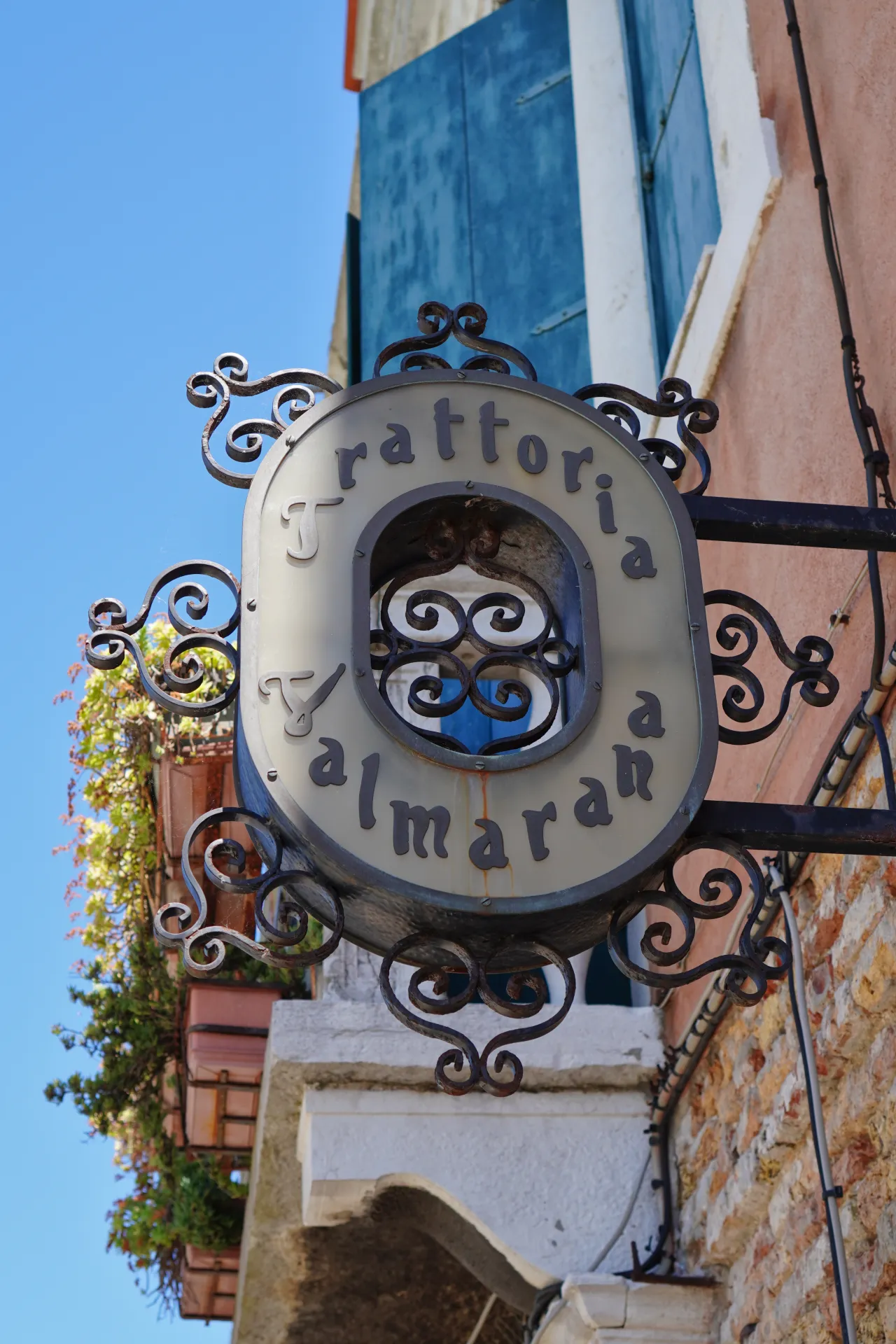 Close-up of the ornate sign for Trattoria Almaran, located on the island of Murano in Venice, Italy, with a clear blue sky and Venetian architecture in the background.