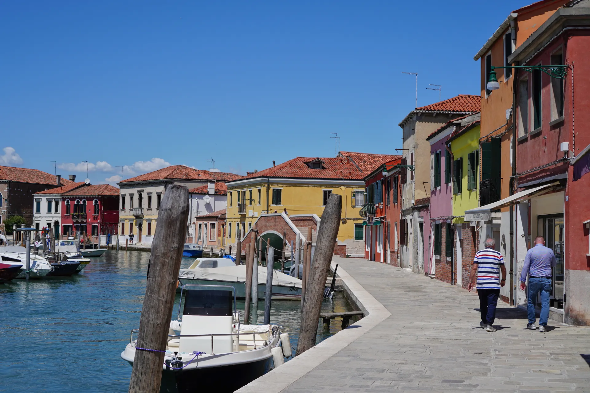 Dva muškarca hodaju popločanom stazom pored kanala na ostrvu Murano, Venecija, sa šarenim zgradama i čamcima na vidiku.