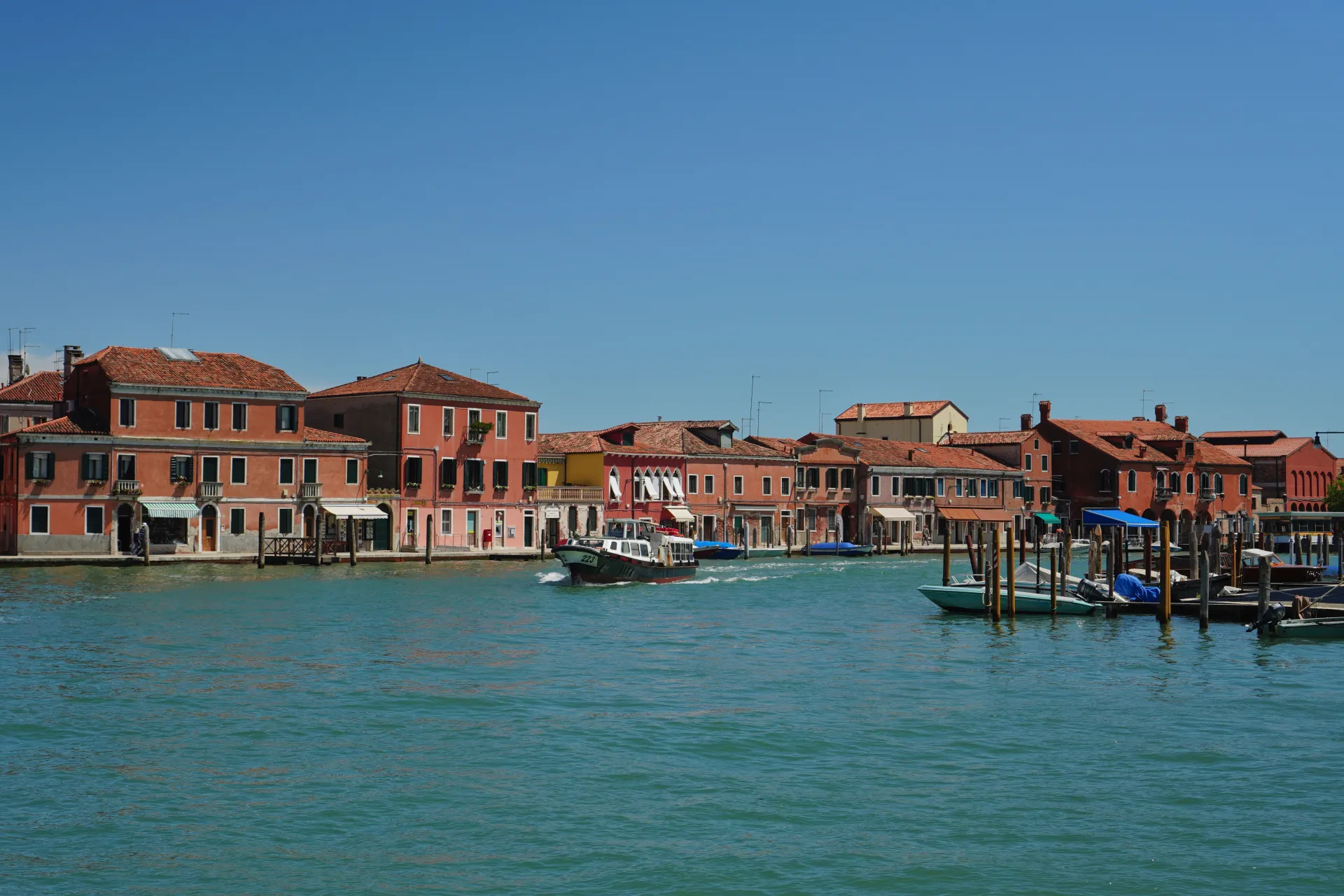 A boat navigates a canal in front of colorful buildings on Murano island, Venice, with docked boats and a bright blue sky overhead.
