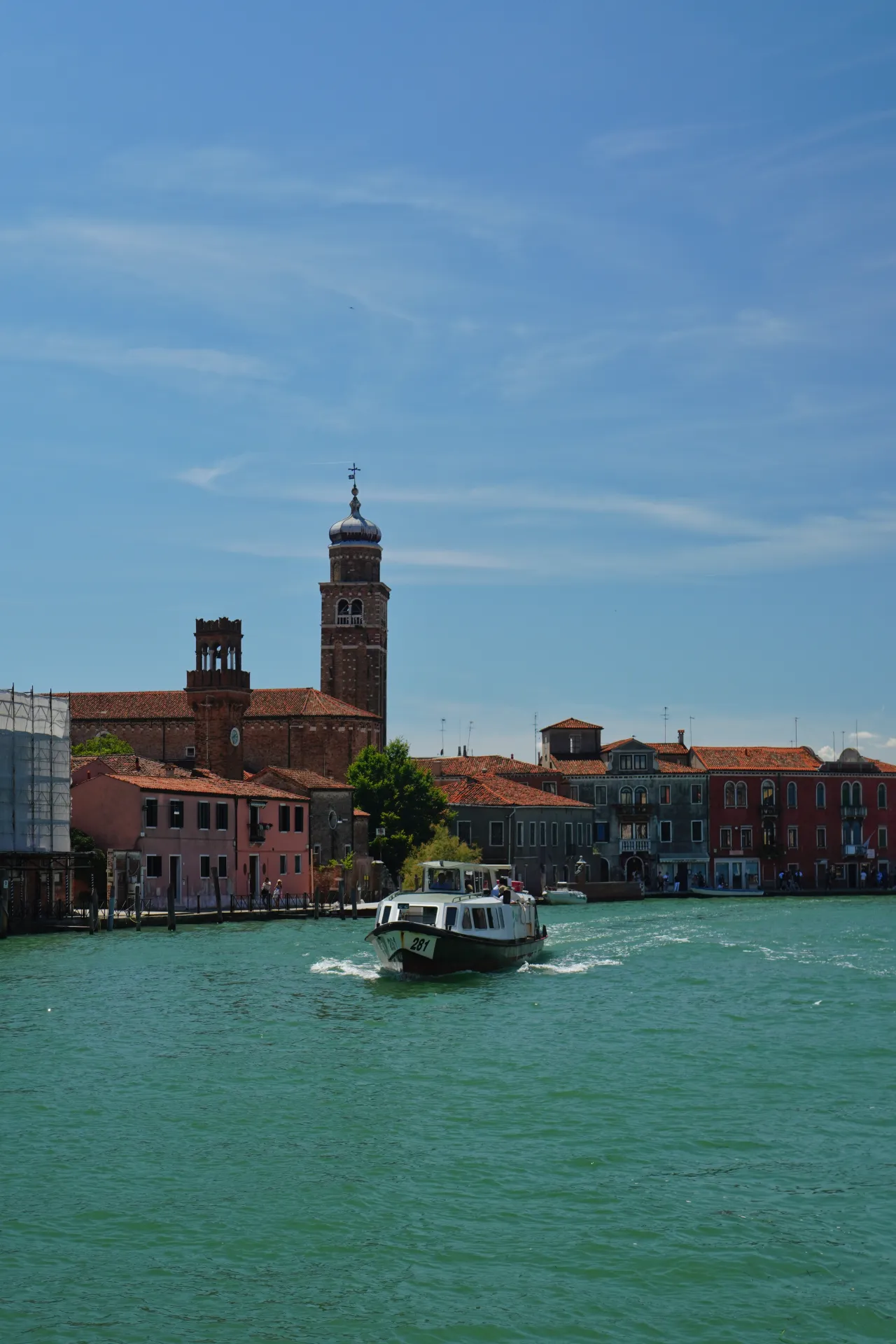 A boat with the number 281 travels on a turquoise canal in Venice, with colorful buildings and a prominent bell tower visible on the island of Murano.
