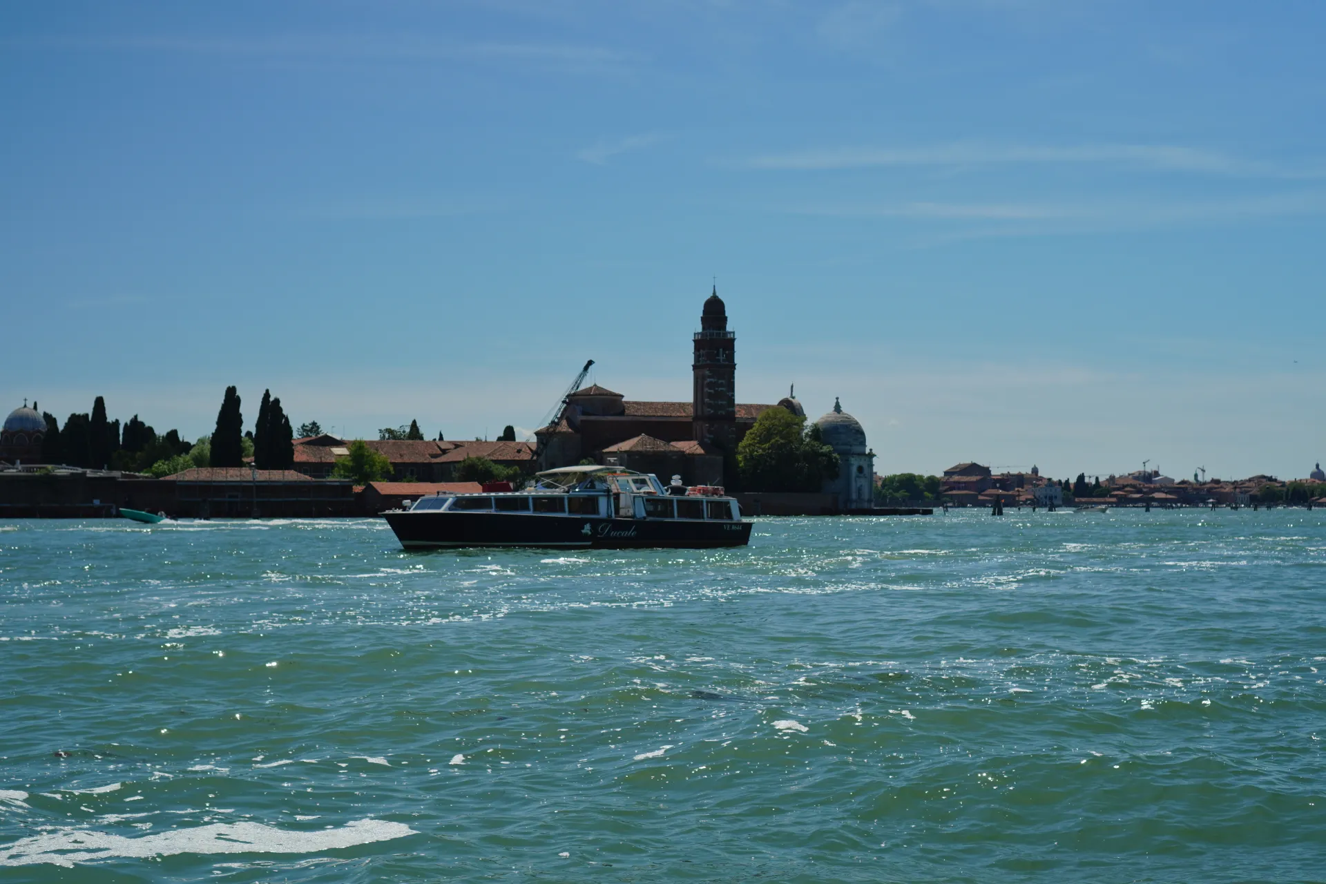 A boat on the turquoise waters of the Venetian lagoon, with the historic buildings and bell towers of Venice visible on the horizon, en route from Murano.