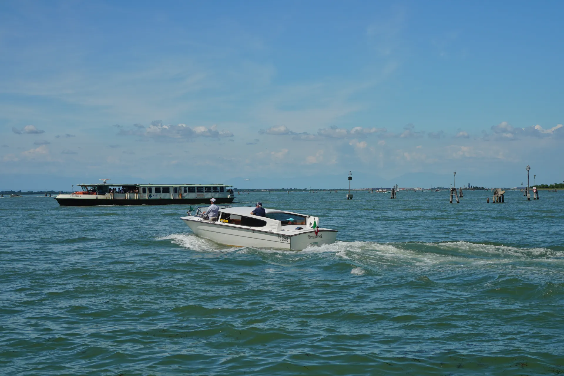 A white motorboat named 'Tomahawk' with two people on board speeds through a canal in Venice, creating a frothy wake. In the background, a larger ferry boat, labeled 'VENEZIA 3', carries passengers. Navigational poles and distant land are visible under a blue sky with scattered clouds.