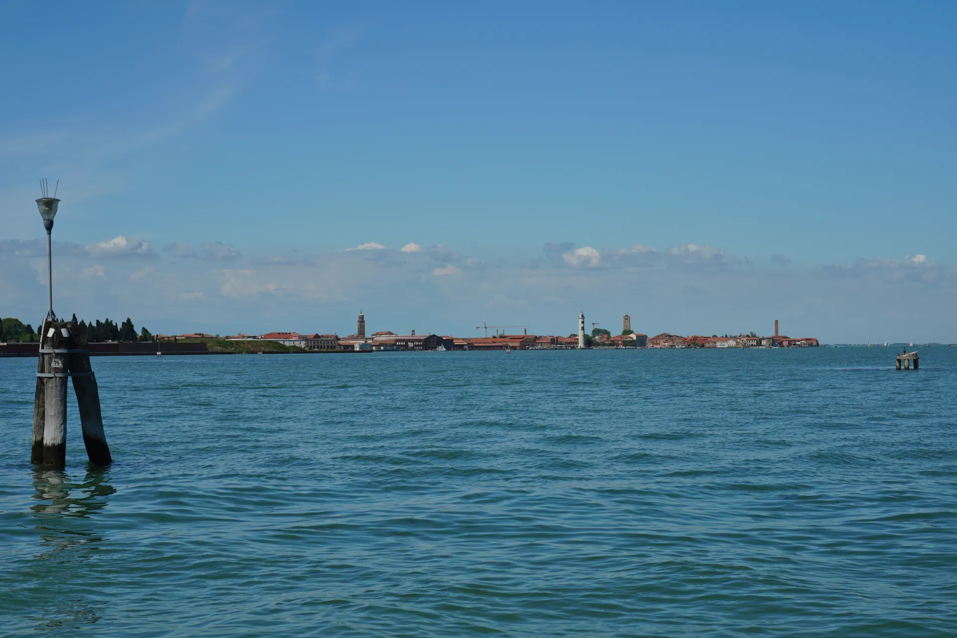 A wide shot of the blue Venetian lagoon with buildings and a lighthouse visible on the horizon, and a wooden post with a light on it in the foreground.