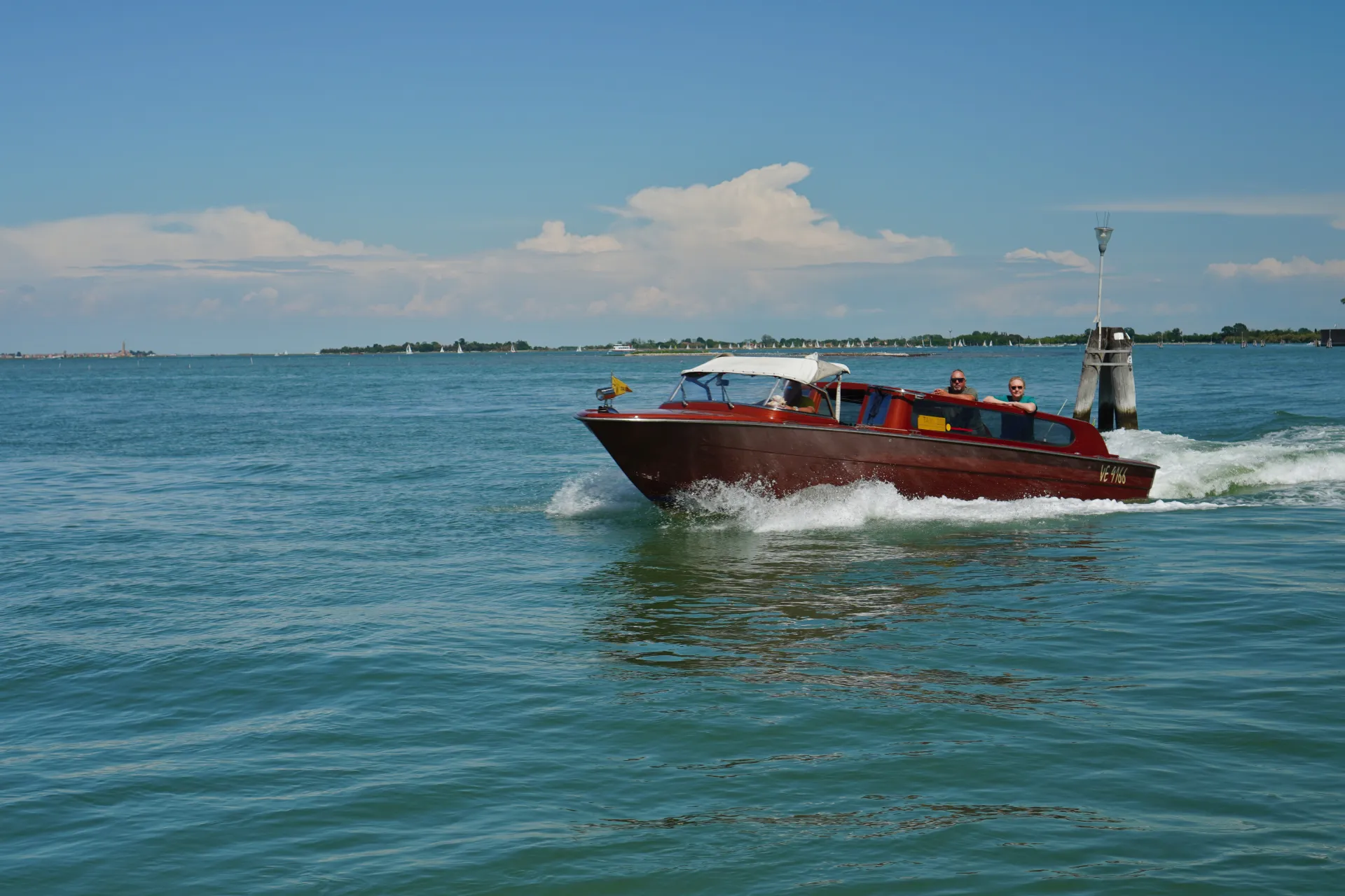 A red wooden Venetian water taxi with two passengers speeds across a bright blue lagoon, leaving a wake behind it. The skyline of Venice and sailboats are visible in the background under a cloudy sky.