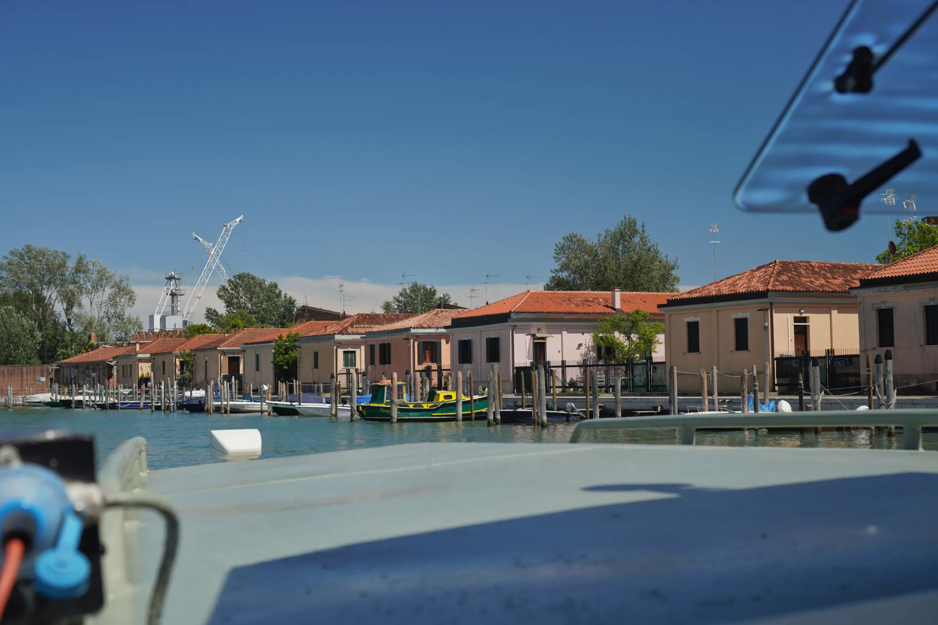 Kanal u Veneciji sa nizom šarenih kuća sa krovovima od crvenog crepa i nekoliko čamaca usidrenih uz vodu, uključujući uočljiv zeleno-žuti čamac. U pozadini se vide građevinski kranovi pod jasnim plavim nebom.