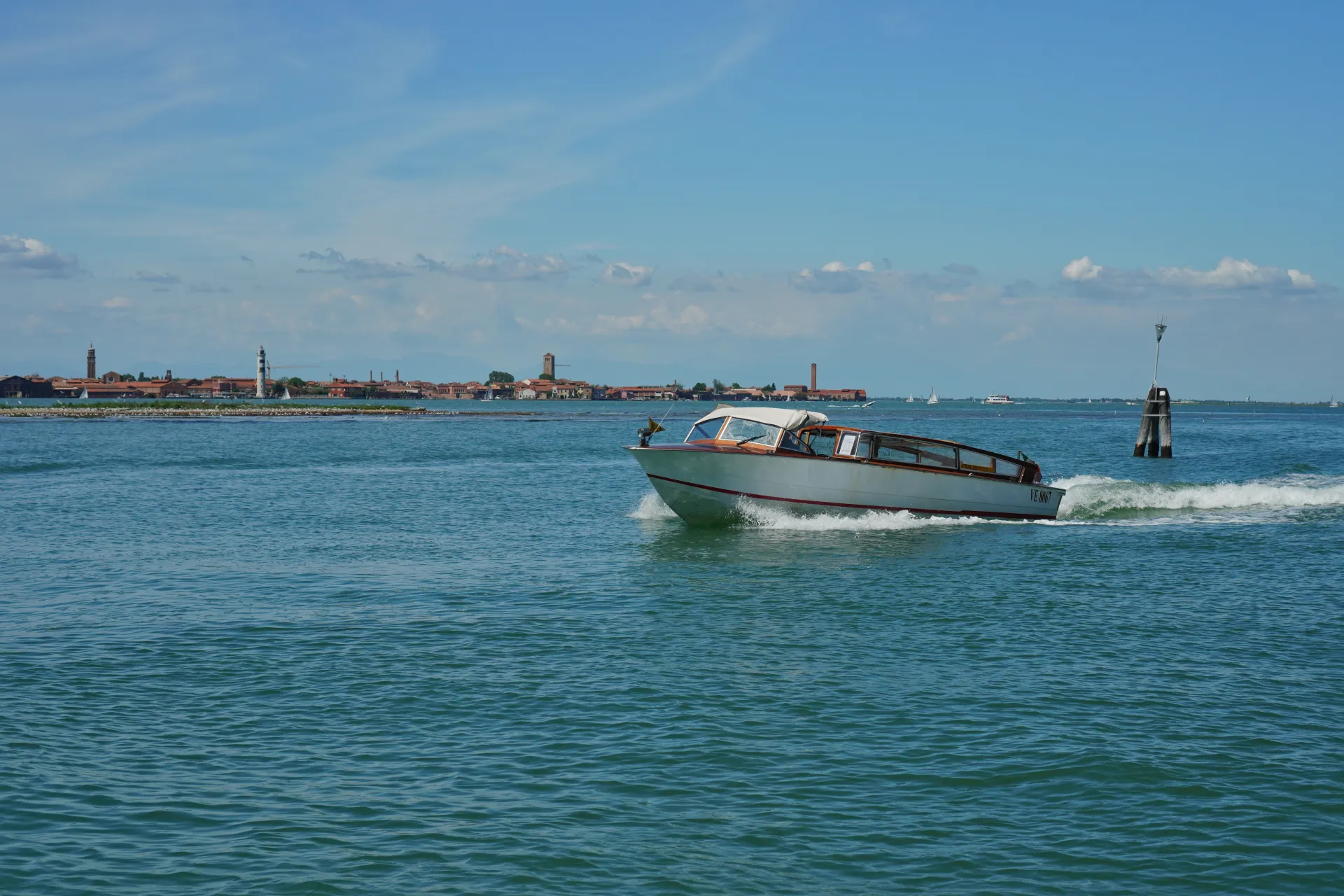 A motorboat cruises through a sunny Venetian canal, with the historic cityscape and lighthouse visible in the distance under a blue sky.