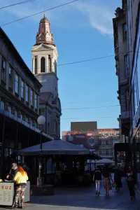 A street in Zagreb, Croatia, with a tall church tower visible above the buildings, and people walking past outdoor cafe seating.