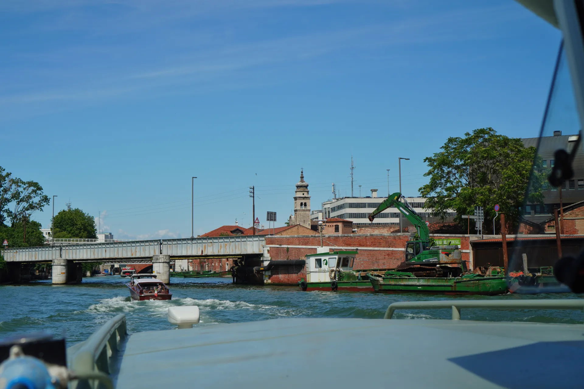 Sunčan dan u Veneciji, čamac plovi kanalom ispod metalnog mosta. Građevinski brodovi i bager usidreni su pored zgrada od cigle na obali kanala.