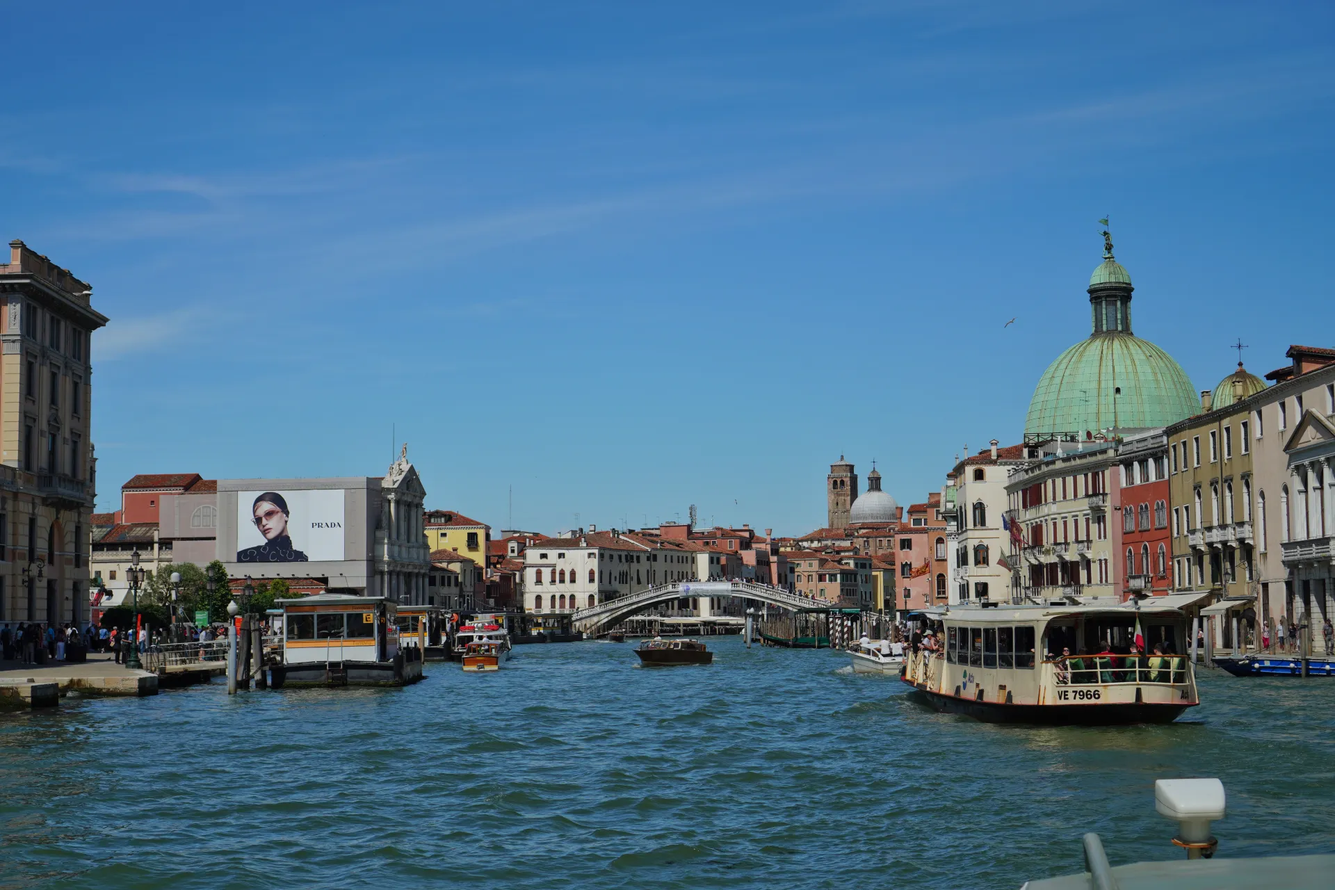 Boats navigating the Grand Canal in Venice, Italy, with colorful buildings and a bridge in the background under a clear blue sky.