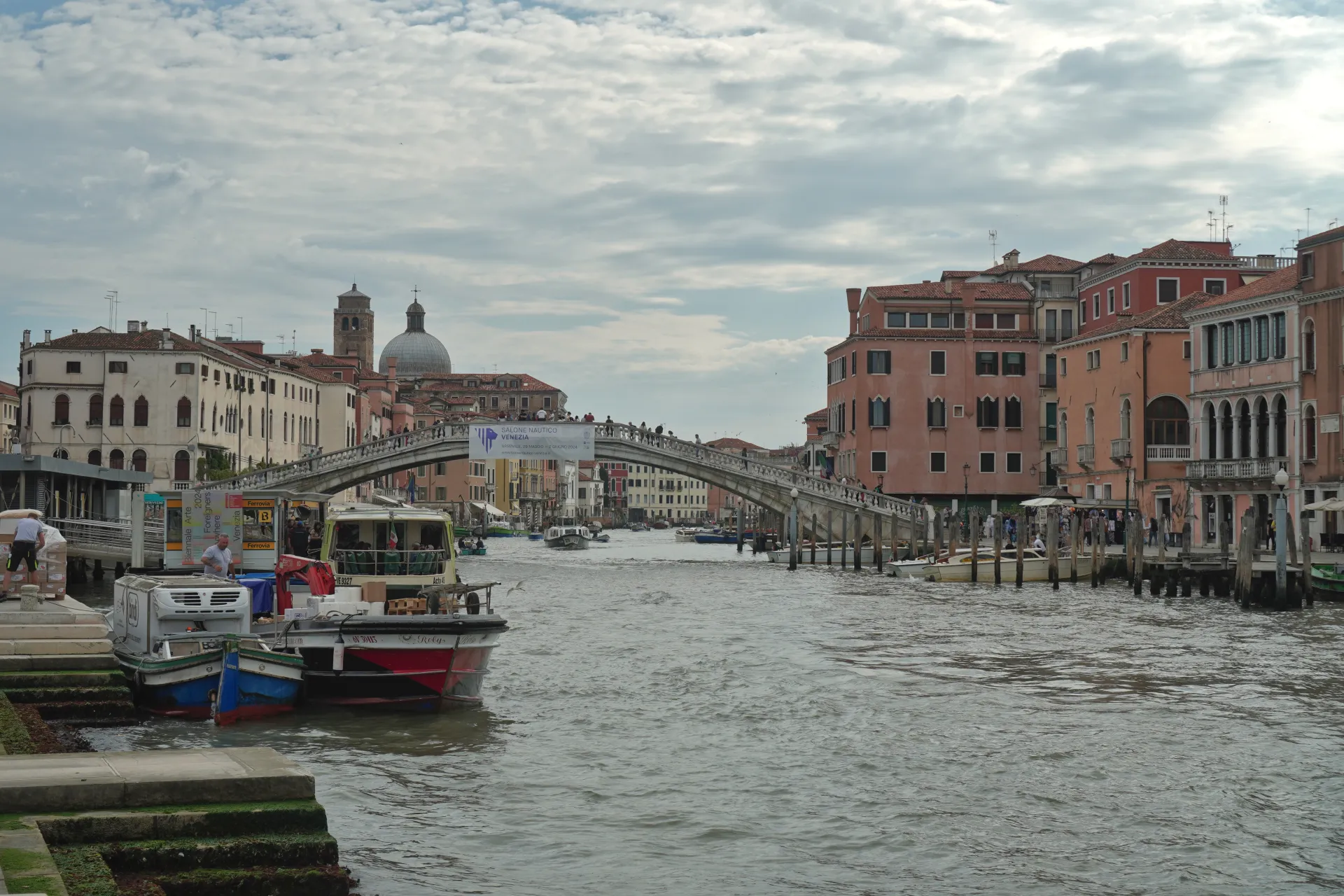 Širok kanal u Veneciji, Italija, sa istaknutim pešačkim mostom koji se izdiže iznad njega. Zgrade sa karakterističnom venecijanskom arhitekturom nižu se uz obale, a na vodi se vide brojni čamci, uključujući trajekt i manje plovile.