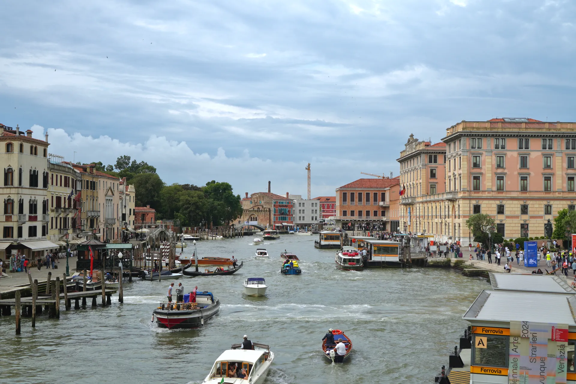 View of the Grand Canal in Venice, Italy, filled with various boats and watercraft, with historic buildings and a cloudy sky in the background.