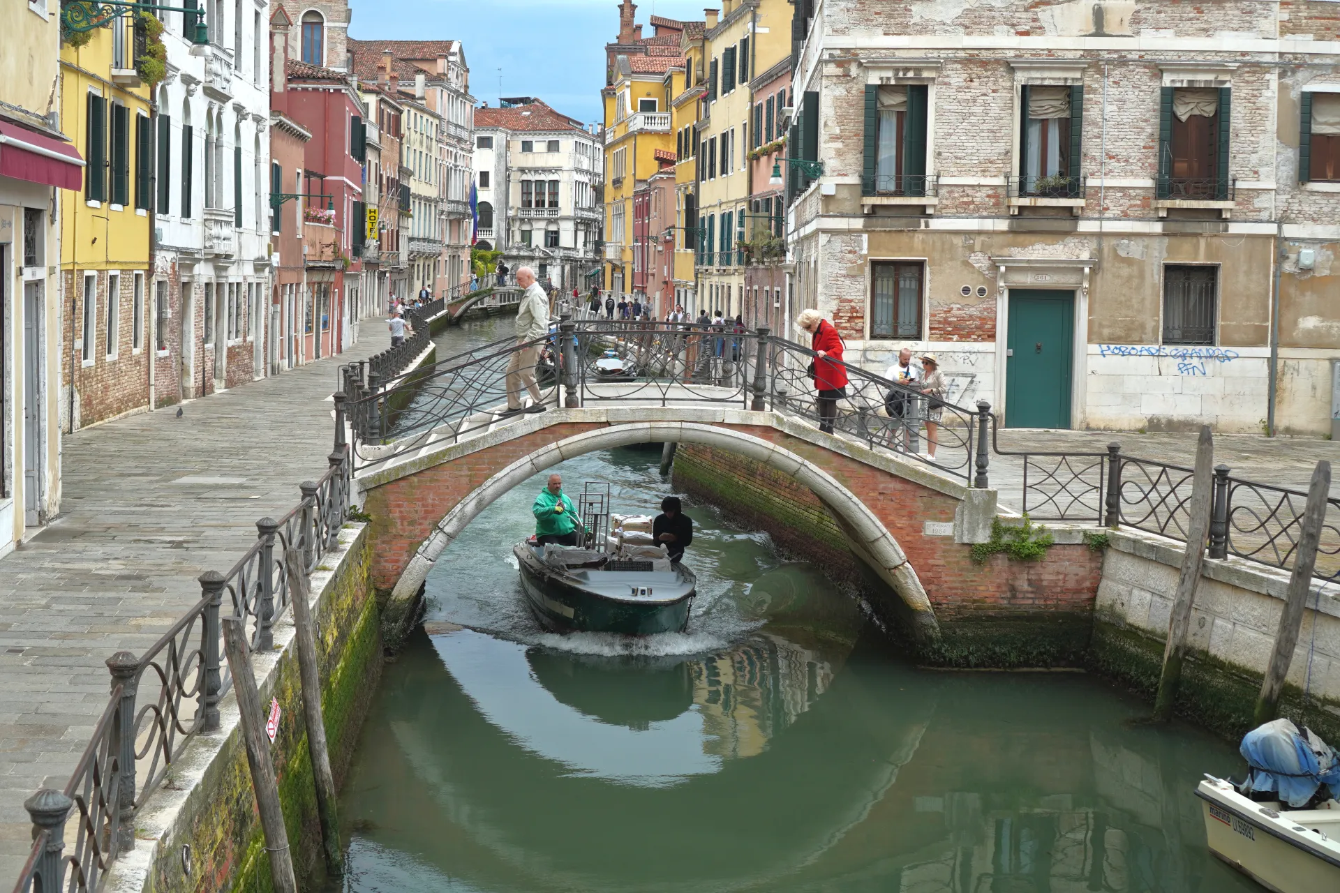 Čamac prolazi ispod ciglanog lučnog mosta iznad kanala u Veneciji, Italija, sa šarenim zgradama duž vodenog puta i ljudima na mostovima.