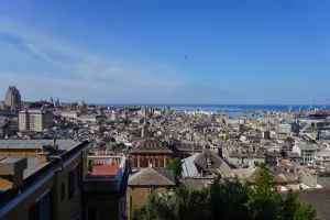 Panoramic view of Genoa, Italy, showing the city's rooftops, church domes, and the harbor with cranes and ships under a clear blue sky.