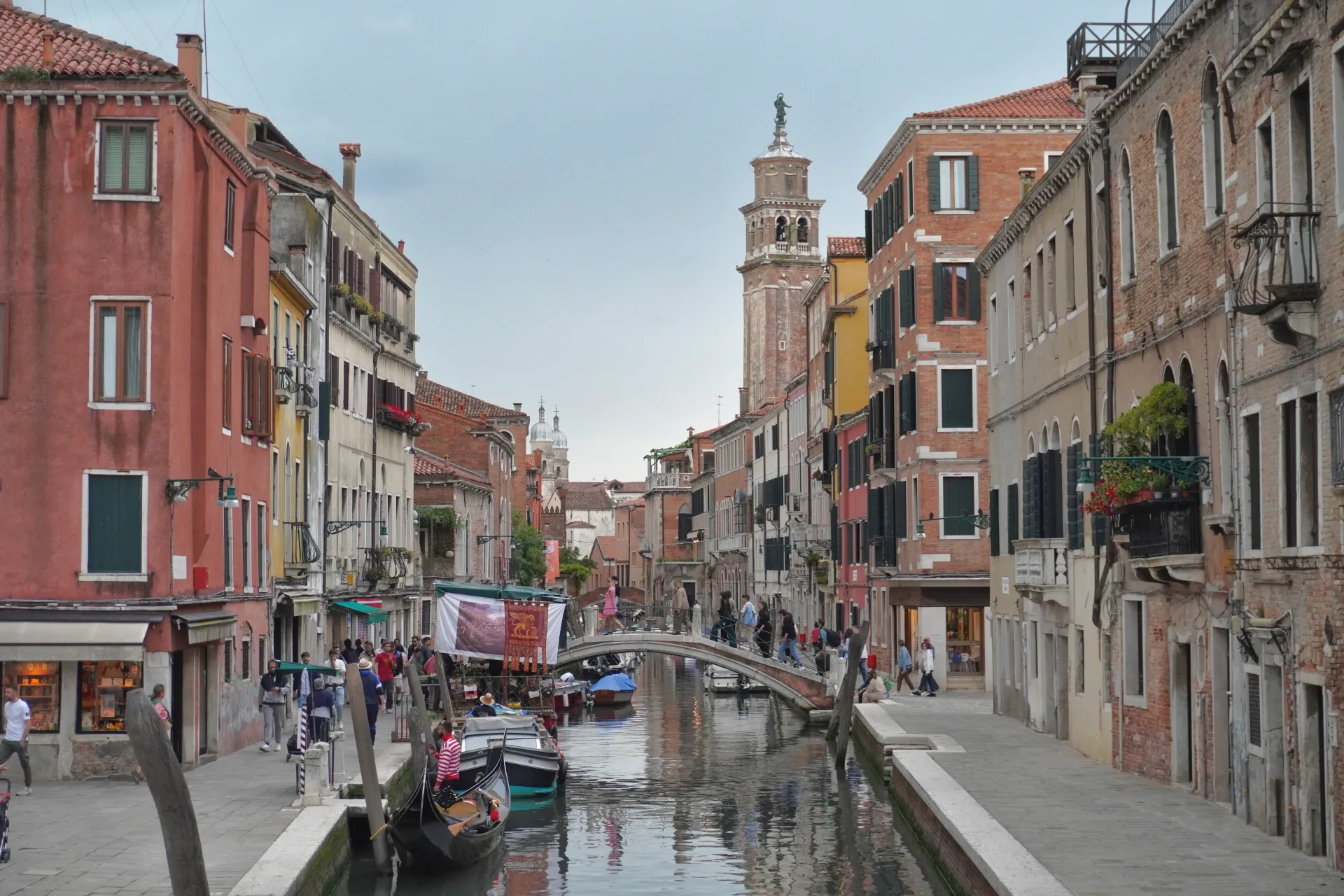 A narrow Venetian canal with buildings on either side, a bridge overhead, and gondolas on the water, with a bell tower in the background.