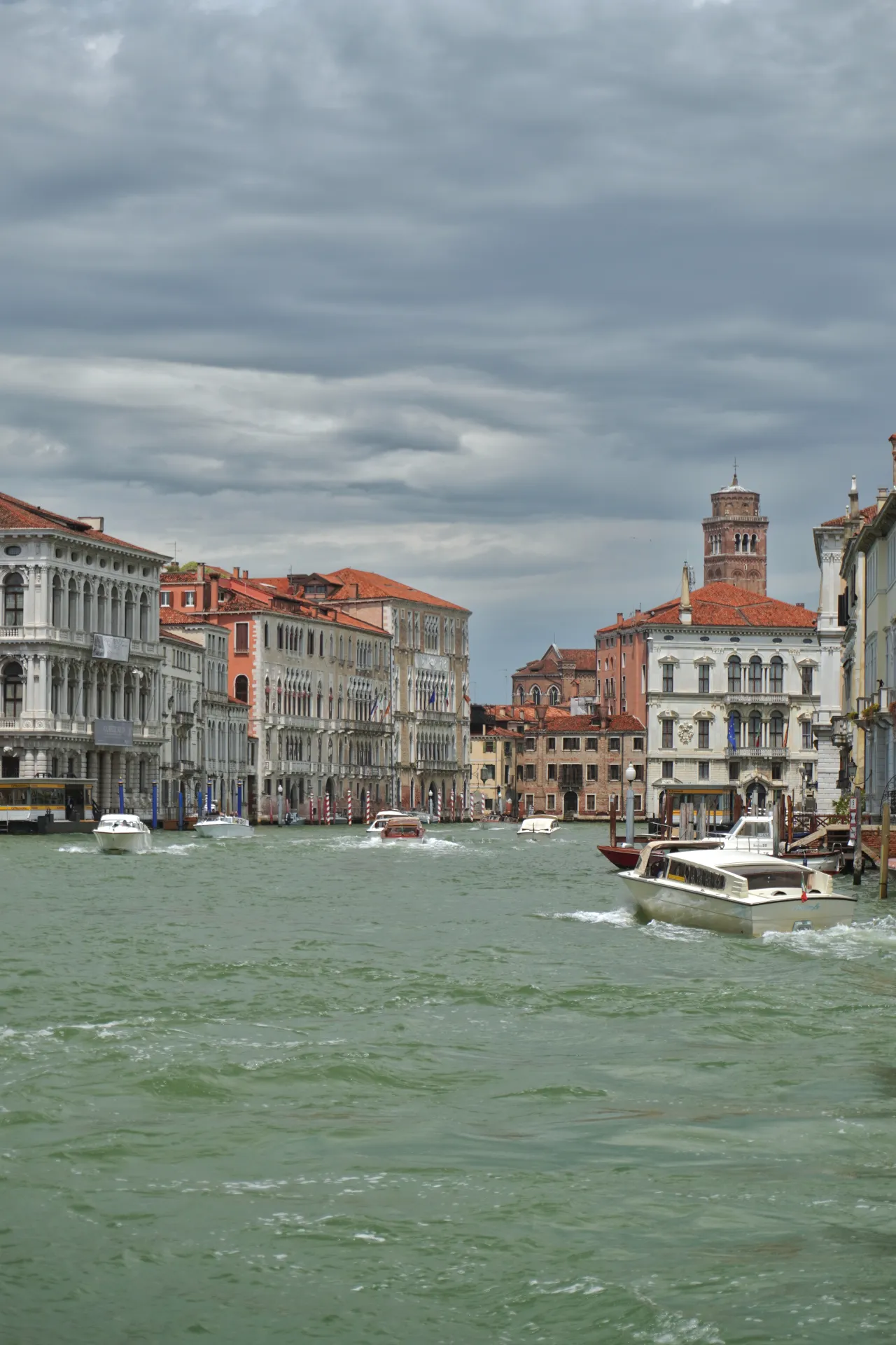 Boats on the Grand Canal in Venice, Italy, with ornate buildings and a bell tower in the background under a cloudy sky.