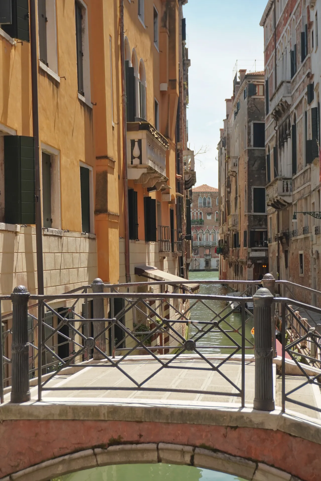 View of a tranquil Venetian canal lined with colorful buildings, looking through the ornate metal railing of a stone bridge.