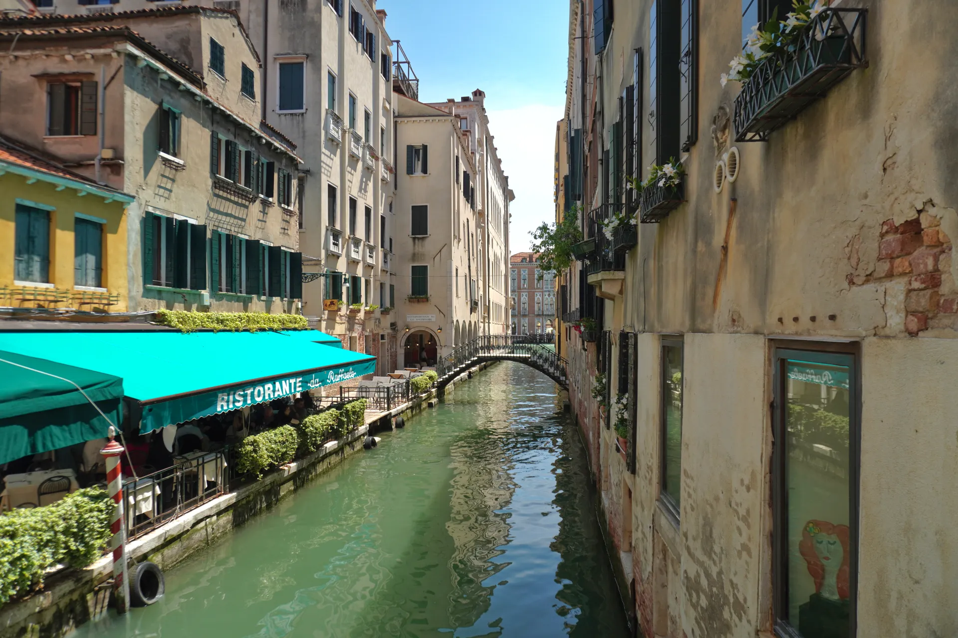 A narrow Venetian canal with traditional buildings on either side, a restaurant with a turquoise awning, and an arched bridge in the background.