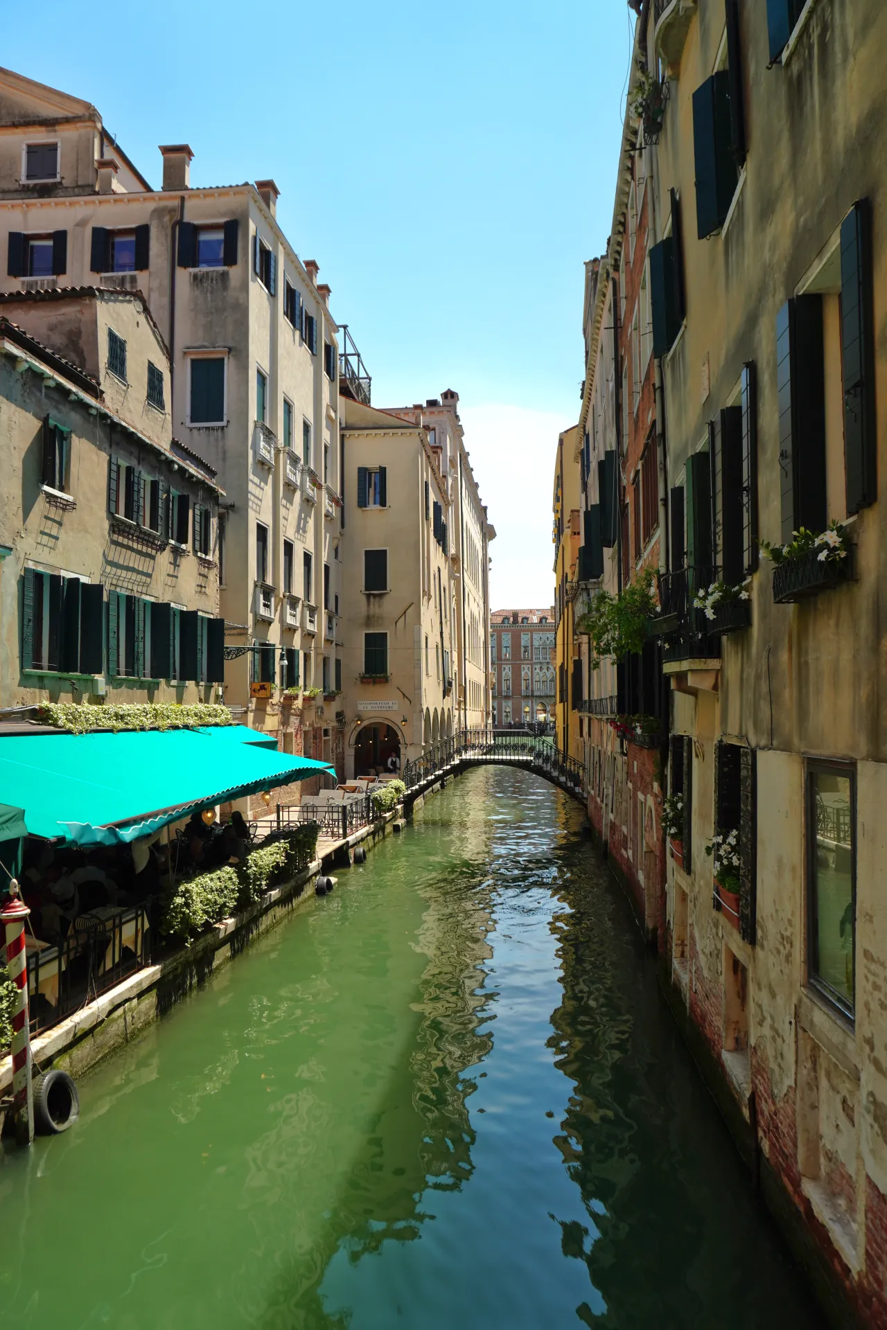Uzak kanal u Veneciji, Italija, sa tradicionalnim zgradama sa obe strane, pešačkim mostom u pozadini i spoljnim kafeom sa zelenom tendom sa leve strane.