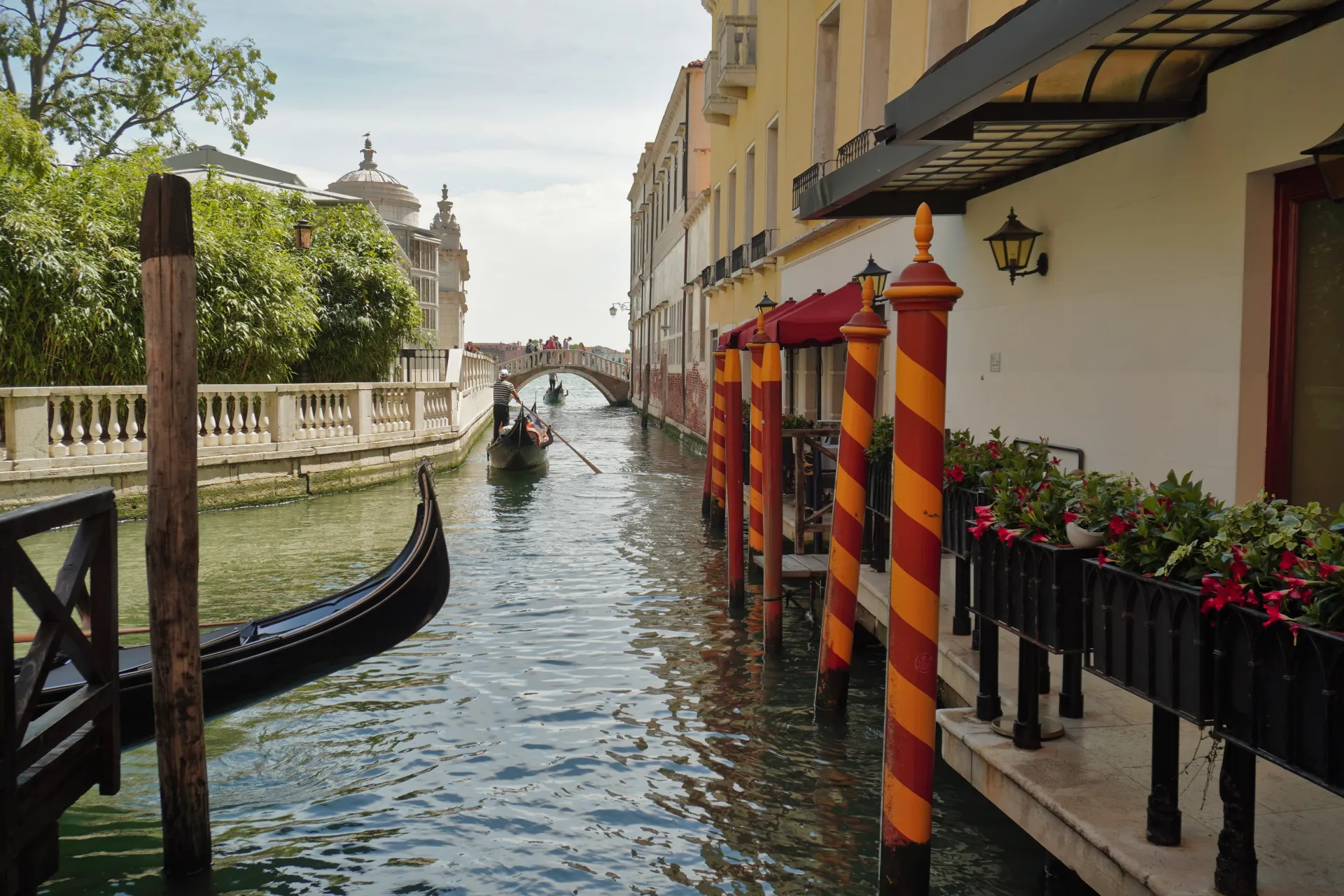 A serene Venetian canal with a gondola in the foreground and another in the distance, passing under a bridge. Traditional Venetian buildings with red awnings and striped mooring poles line the canal.