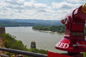 A wide shot showcasing the Danube River with a distant city skyline of Novi Sad, Serbia, viewed from a high vantage point, possibly Petrovaradin Fortress, with a prominent red tourist telescope in the foreground.