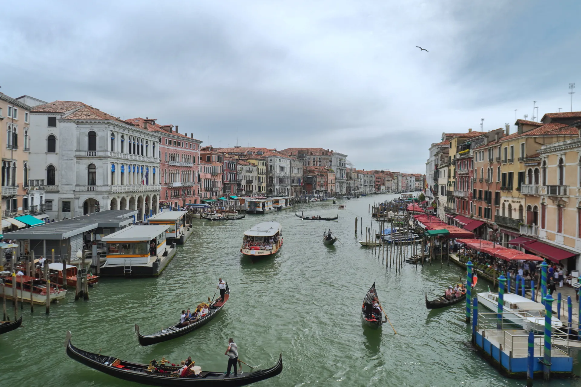 A wide view of the Grand Canal in Venice, Italy, featuring several gondolas with passengers, a water bus, and ornate historic buildings on both sides of the canal under a cloudy sky.