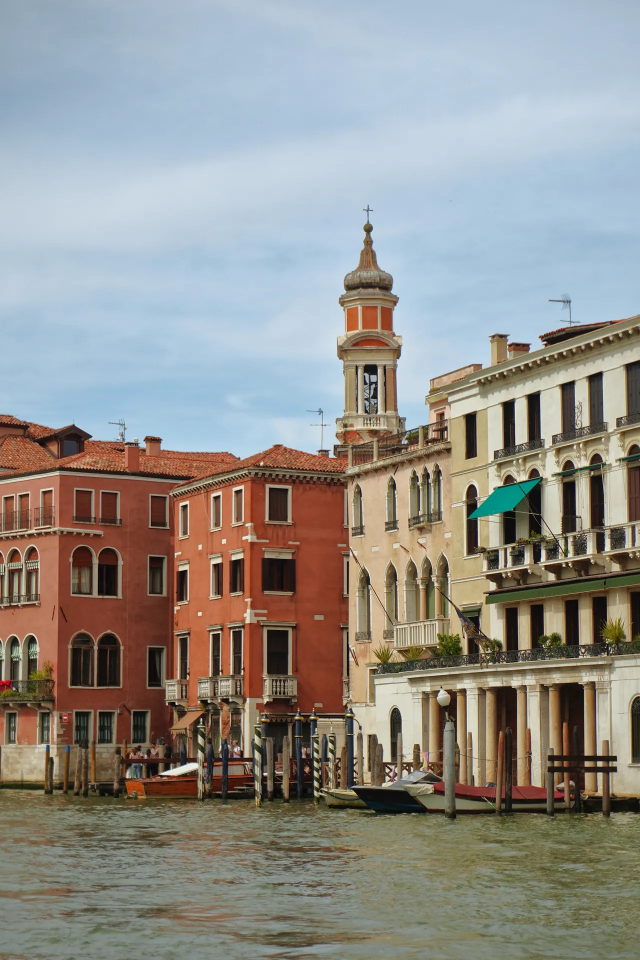Colorful Venetian buildings with a bell tower overlook the Grand Canal, with several boats docked along the waterfront.