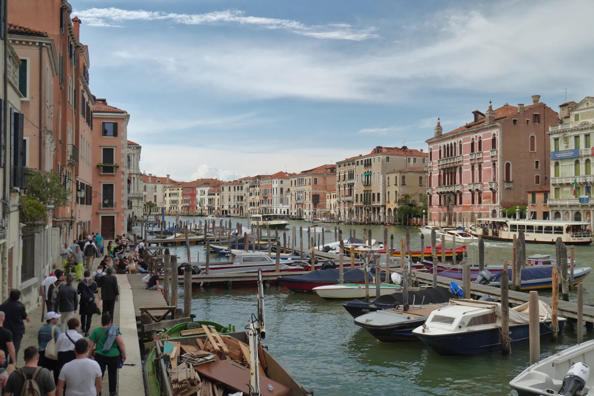 A wide canal in Venice, Italy, with numerous docked boats and vaporettos. Historic buildings line the canal, and people are walking on the promenade.