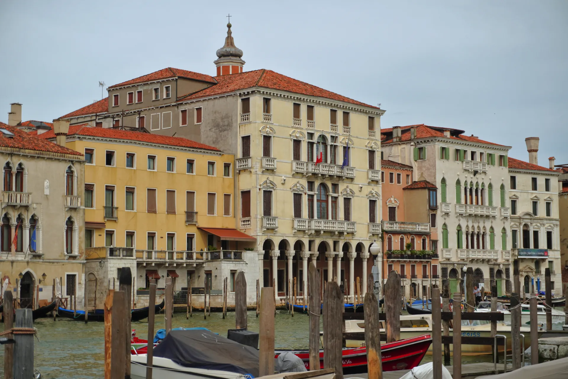 A wide canal in Venice, Italy, with several gondolas and motorboats moored along wooden posts. Lined with traditional Venetian buildings in shades of yellow, cream, and peach, featuring red tile roofs and ornate architectural details.