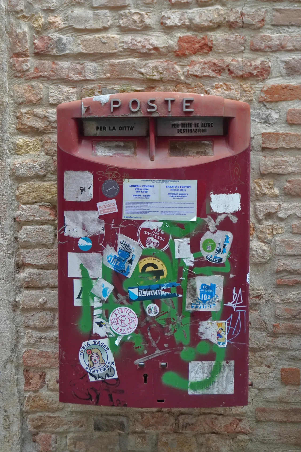 Красный итальянский почтовый ящик с надписью 'POSTE', покрытый различными наклейками и зелеными граффити, закрепленный на обшарпанной кирпичной стене в Венеции.